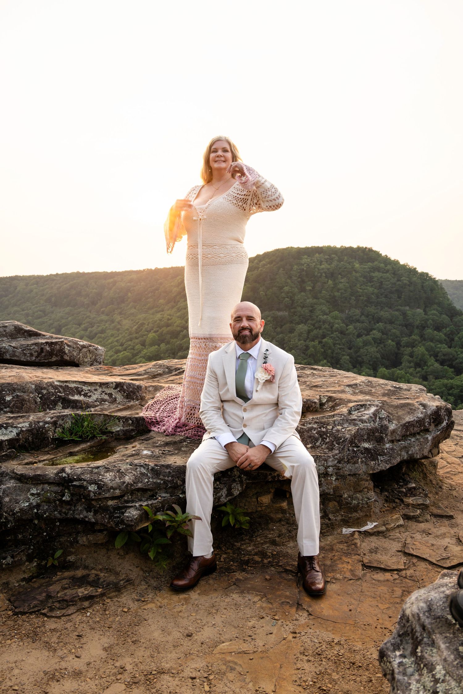 Bride and groom posing on rocky overlook at sunset with lush green mountain valley backdrop.