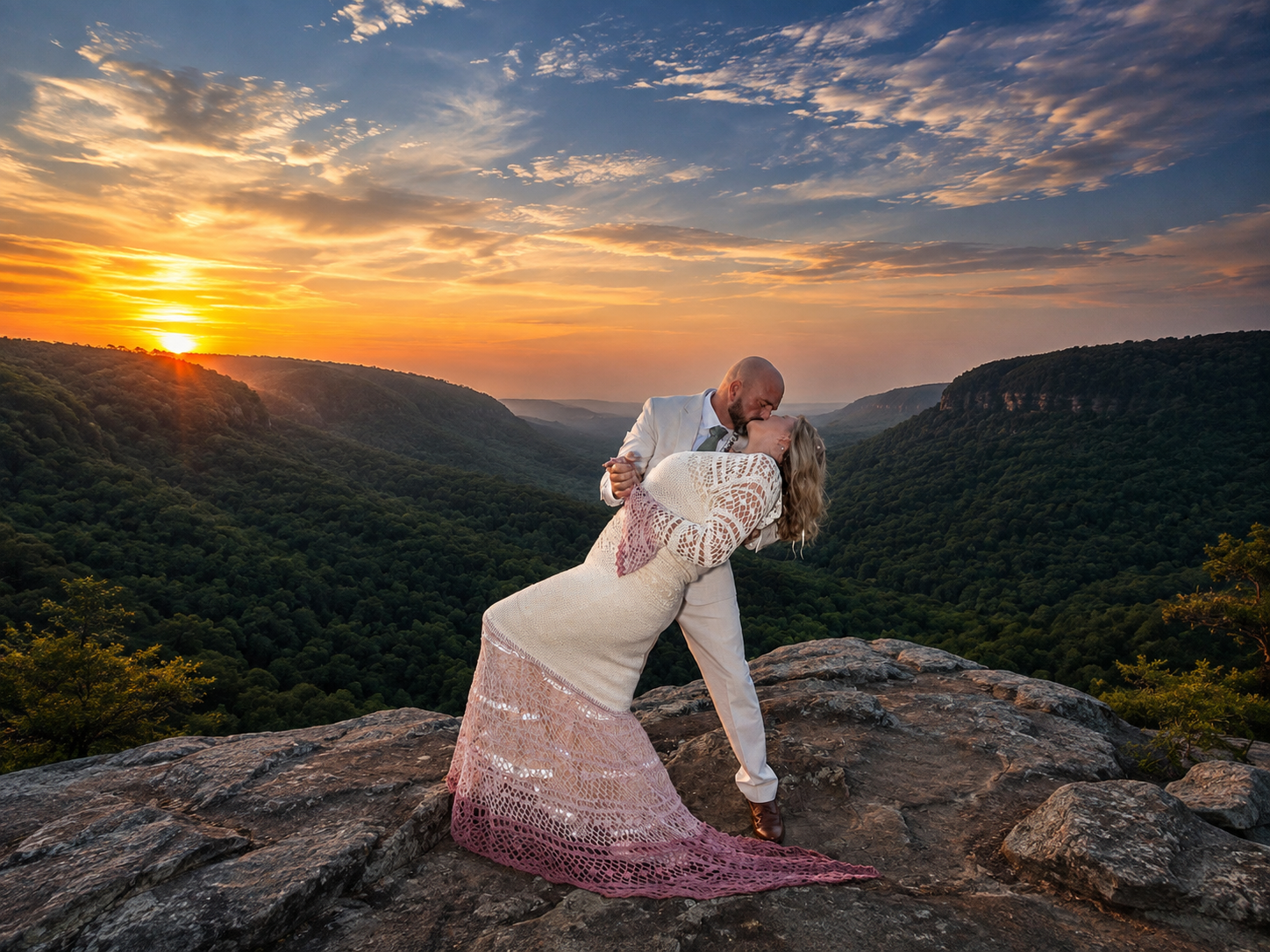 Couple sharing romantic dip kiss on mountain overlook at sunset, bride in white lace gown with ombre train.