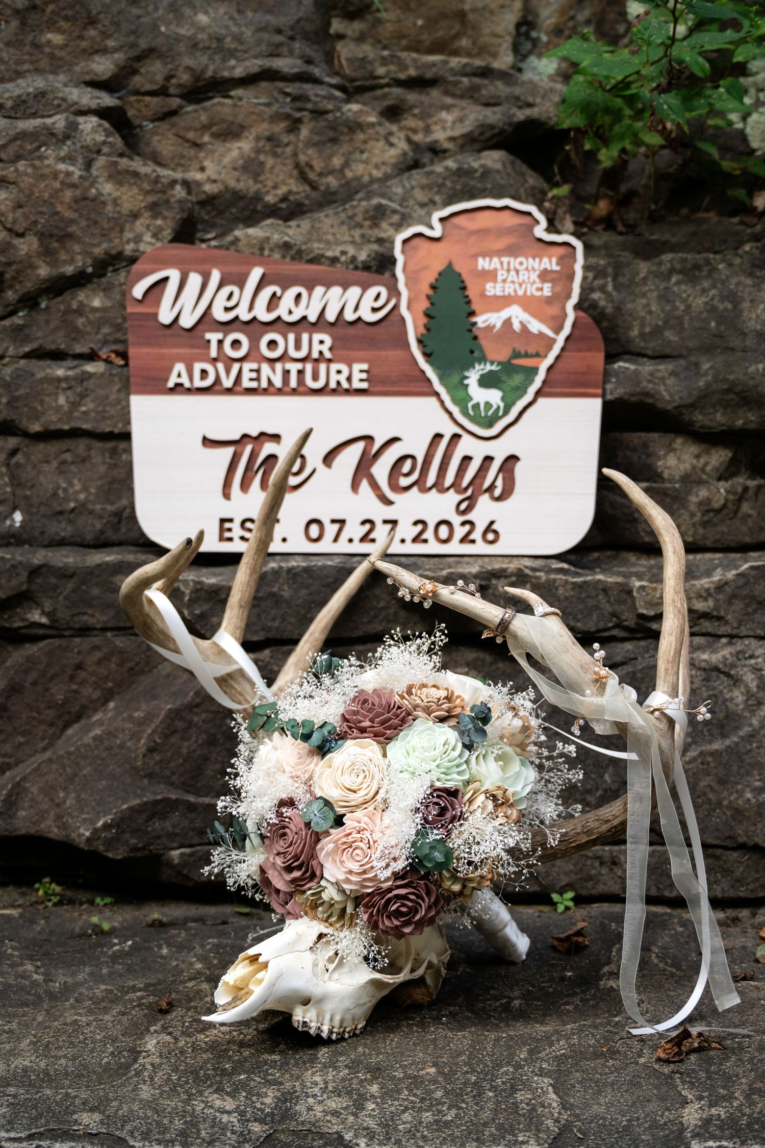 Rustic wedding welcome sign for The Kellys Est. 07.27.2026 with National Park theme, antlers, and floral bouquet on stone wall.