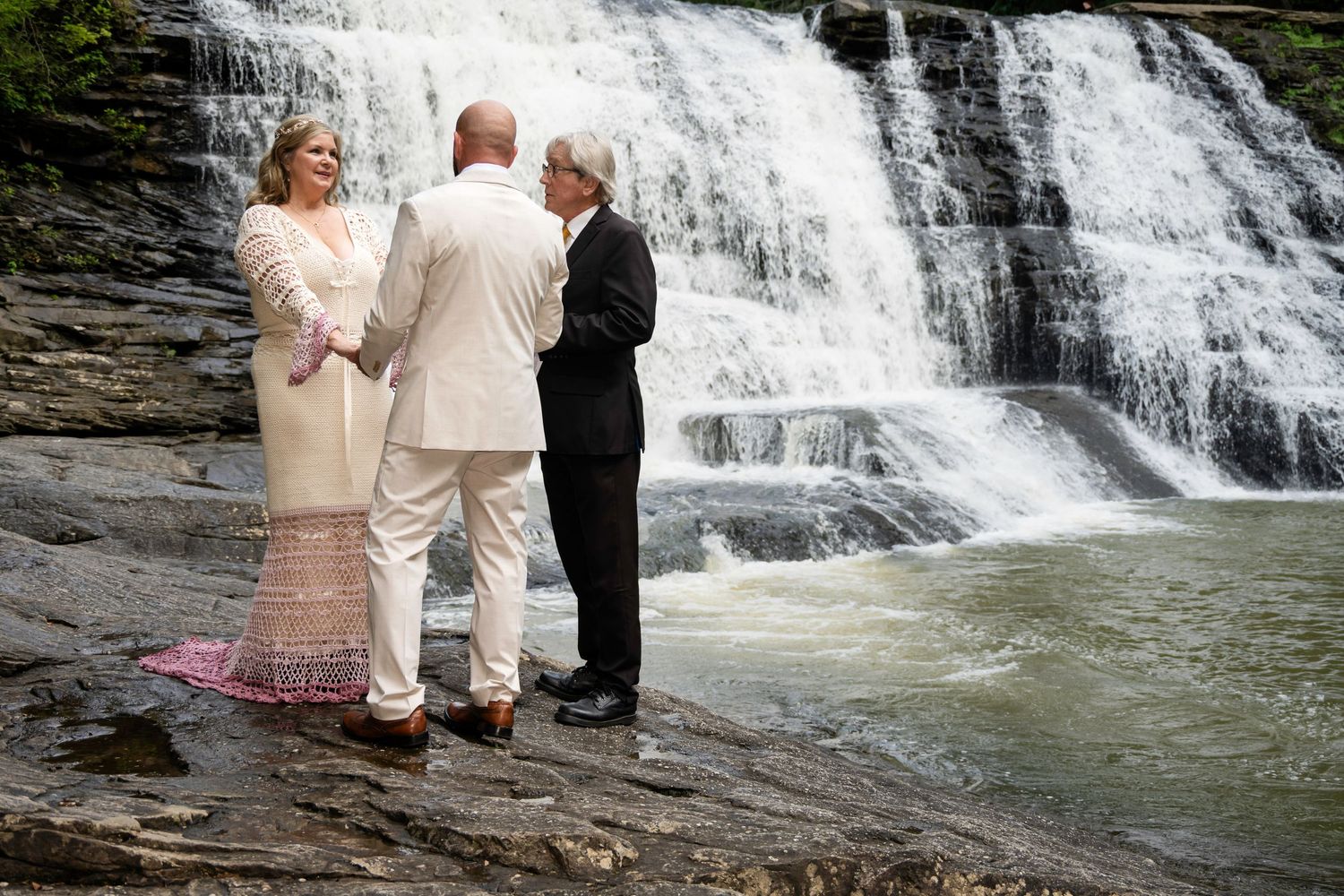 Couple exchanges vows before a stunning waterfall backdrop with an officiant during an outdoor wedding ceremony.