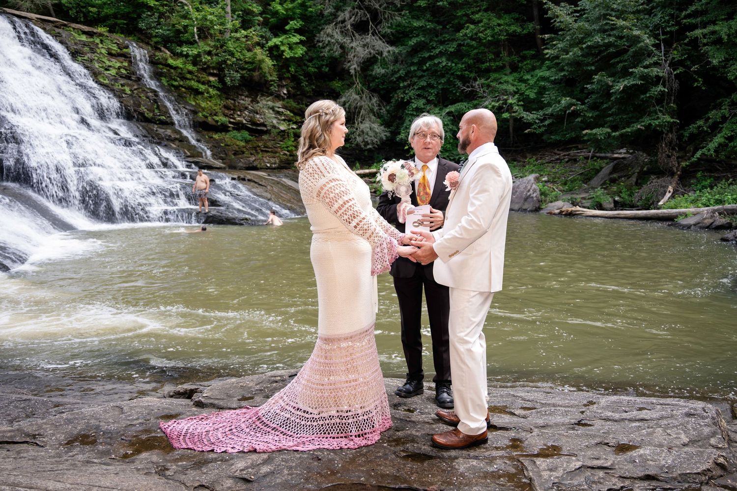 Couple exchanges vows before a waterfall with an officiant, bride in lace gown, groom in white suit, outdoor wedding.