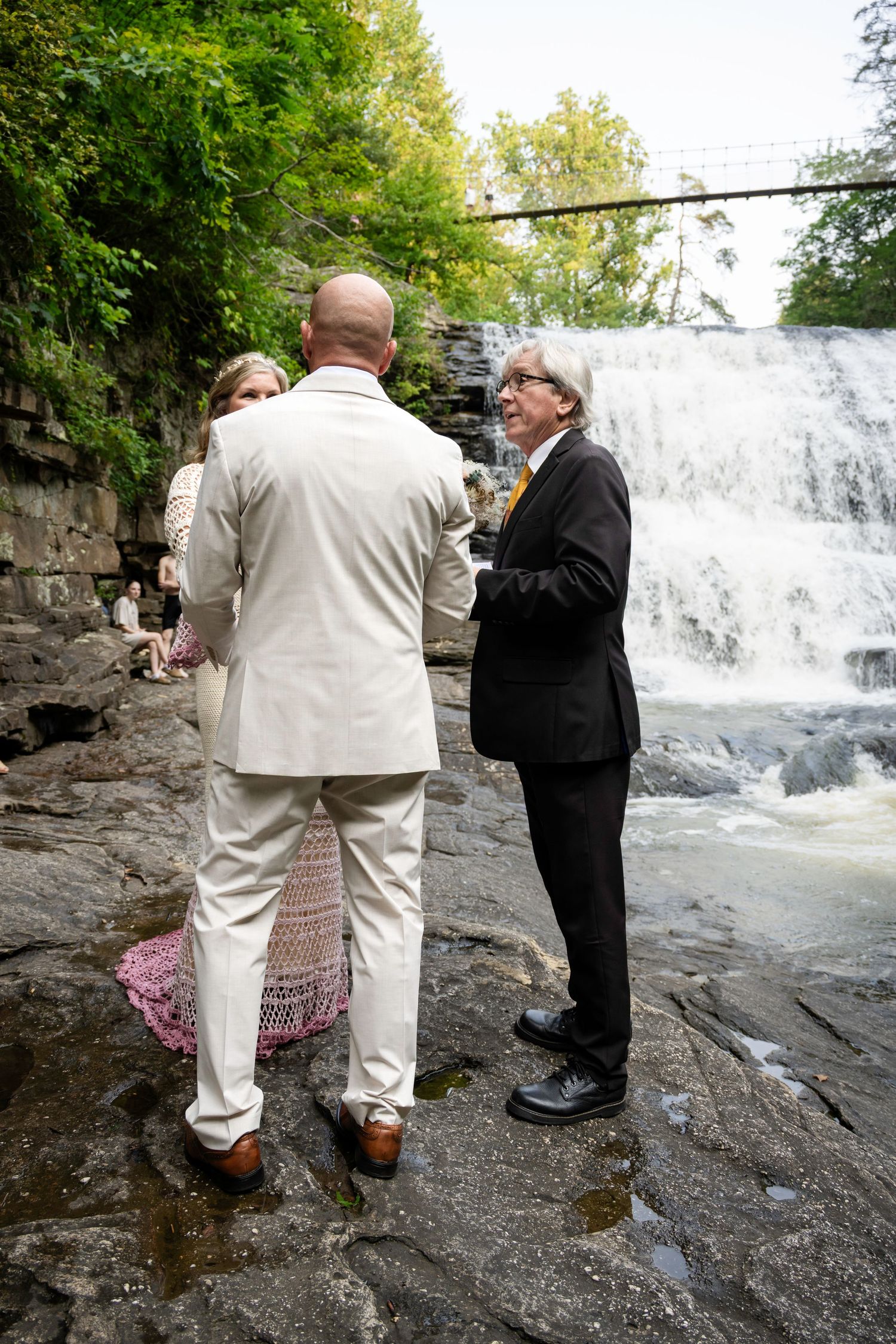 Couple exchanging vows with officiant beside a stunning waterfall on rocky terrain outdoors.
