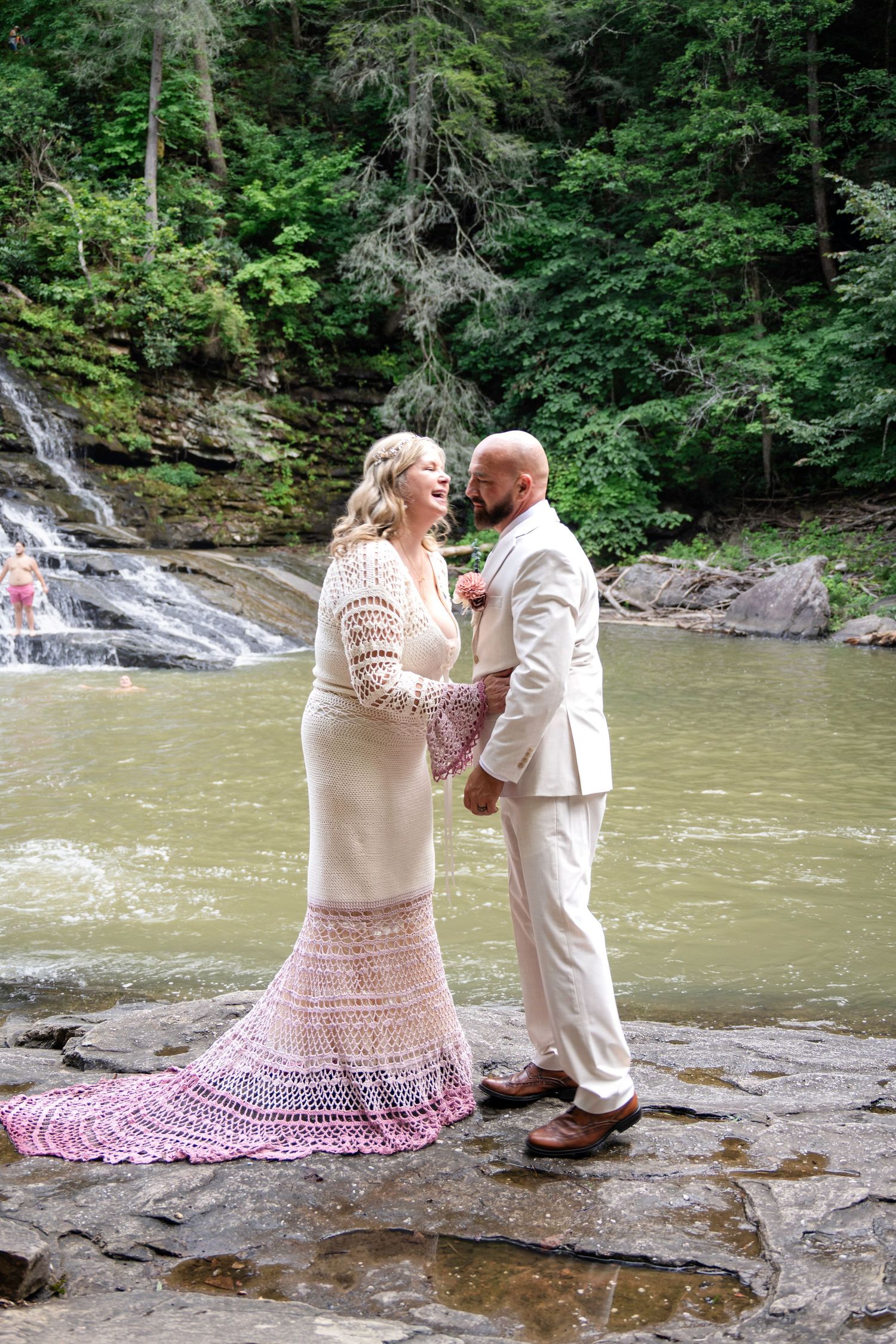 Bride and groom stand on rocks before a waterfall, she in a pink crochet gown, he in a cream suit.
