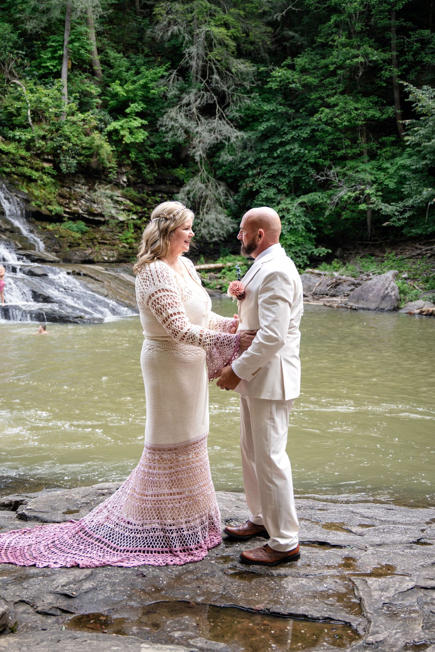 Couple exchanges vows on rocky riverbank in white crochet gown and tan suit with waterfall backdrop.