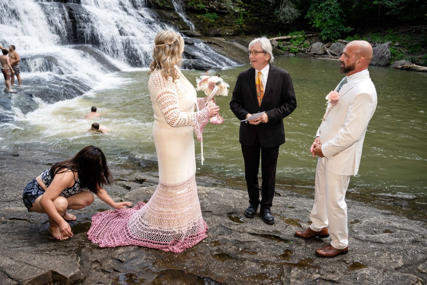Outdoor waterfall wedding ceremony with bride in white gown, groom in cream suit, officiant, and guest on rocky riverbank.