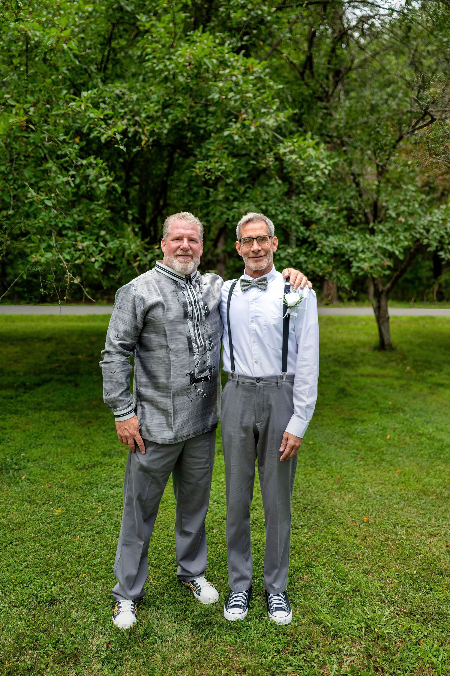 Two men in matching gray suits pose together outdoors on green grass with lush trees in the background.