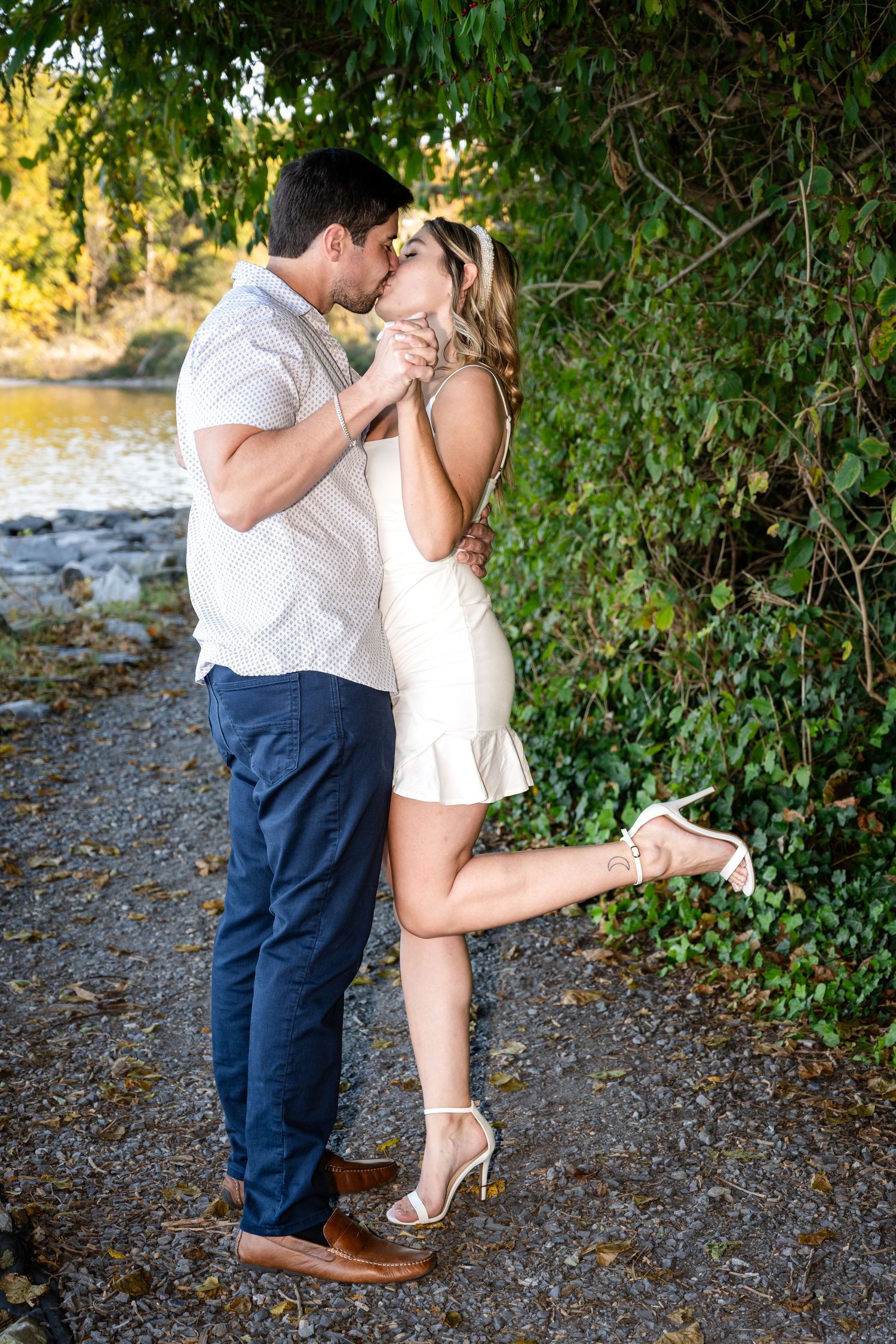 Couple sharing a romantic kiss outdoors near a river, woman in white dress lifting one leg, surrounded by lush green foliage.