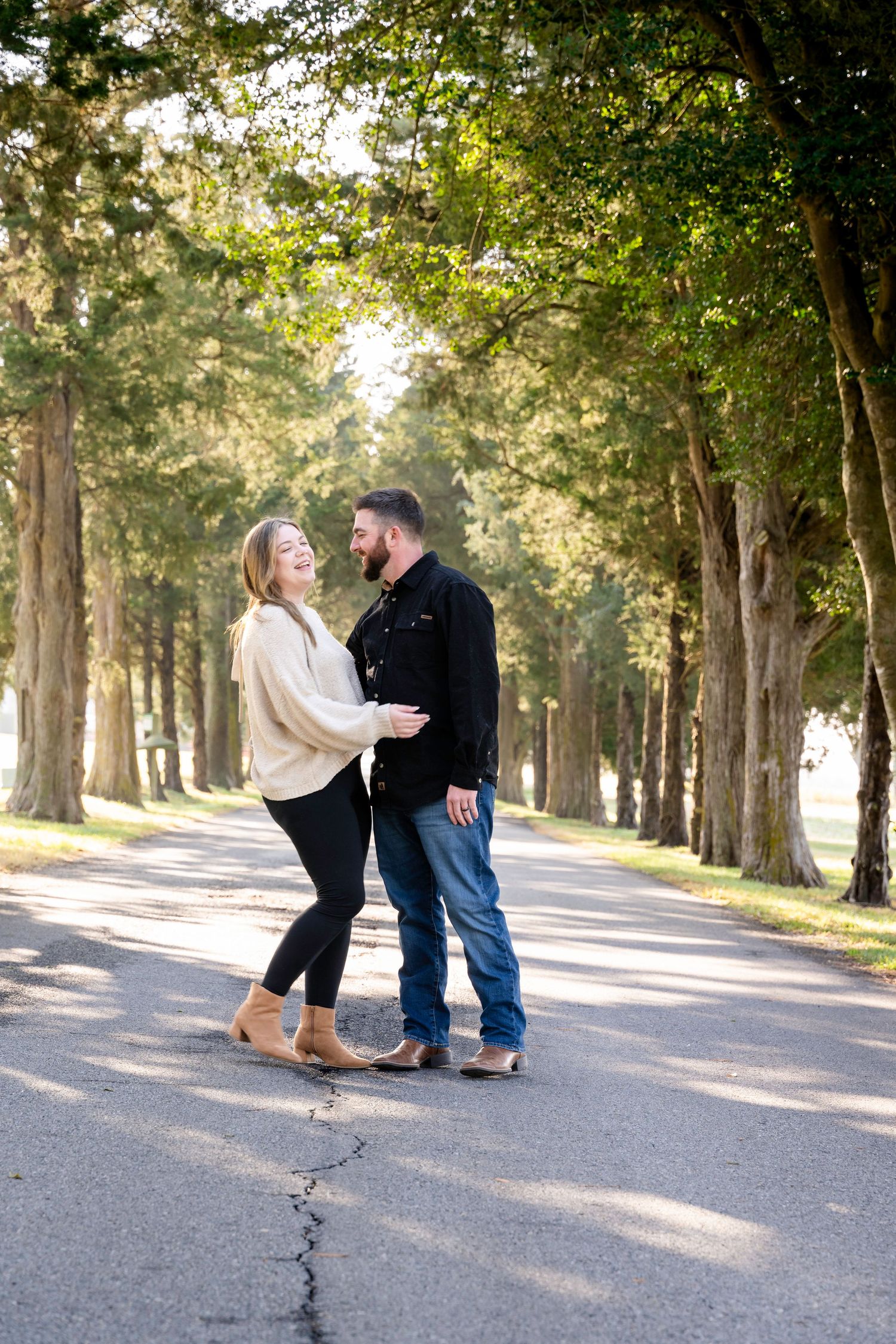 Couple laughing together on a tree-lined path during an outdoor engagement photo session in natural light.