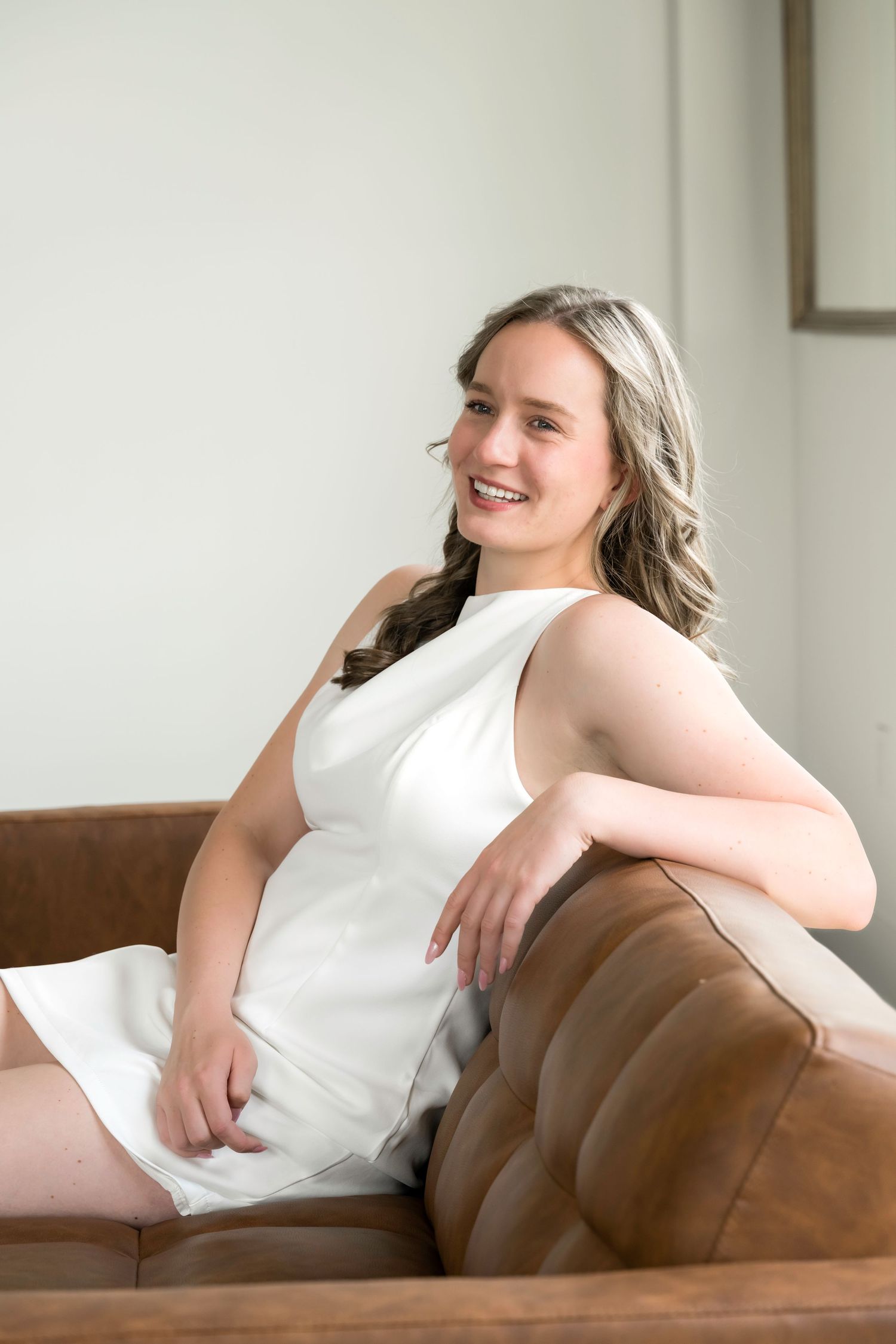 Young woman in white dress smiling while seated on brown leather sofa in bright modern room.