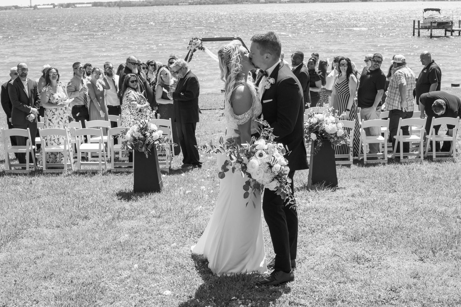 Black and white photo of newlyweds sharing first kiss at outdoor wedding ceremony with guests seated on white chairs.