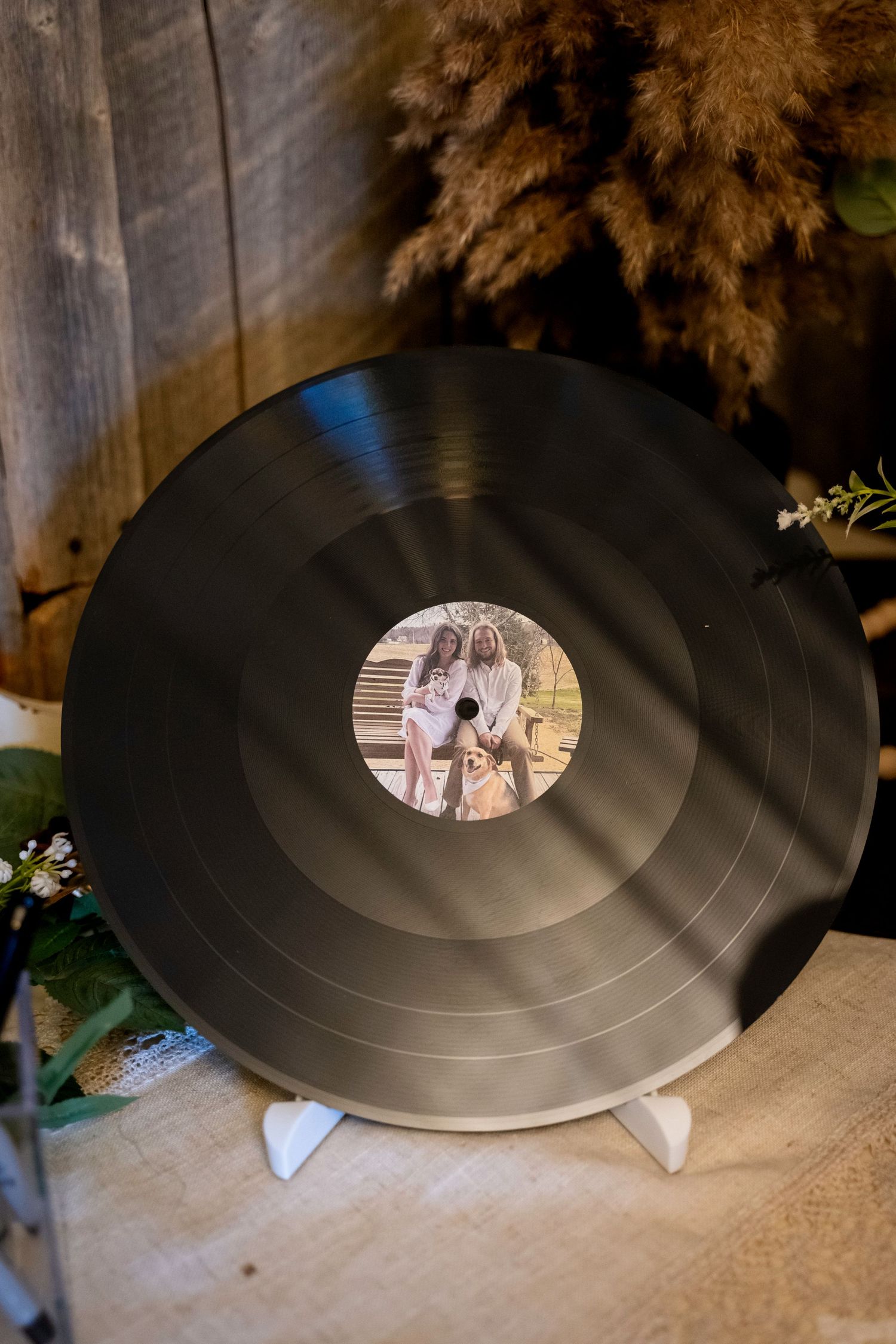 Custom vinyl record wedding keepsake featuring couple photo label displayed on white stand with rustic wooden backdrop.