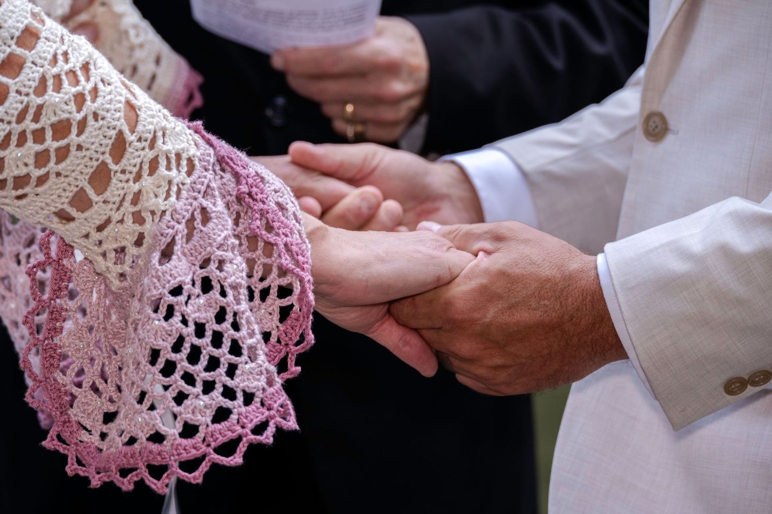 Close-up of a couple exchanging wedding rings during an outdoor ceremony, bride wearing pink lace dress.
