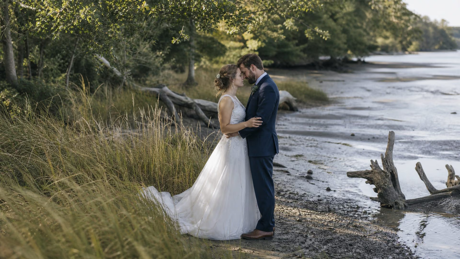 Bride and groom share a romantic moment by a serene riverside, surrounded by lush greenery on their wedding day.