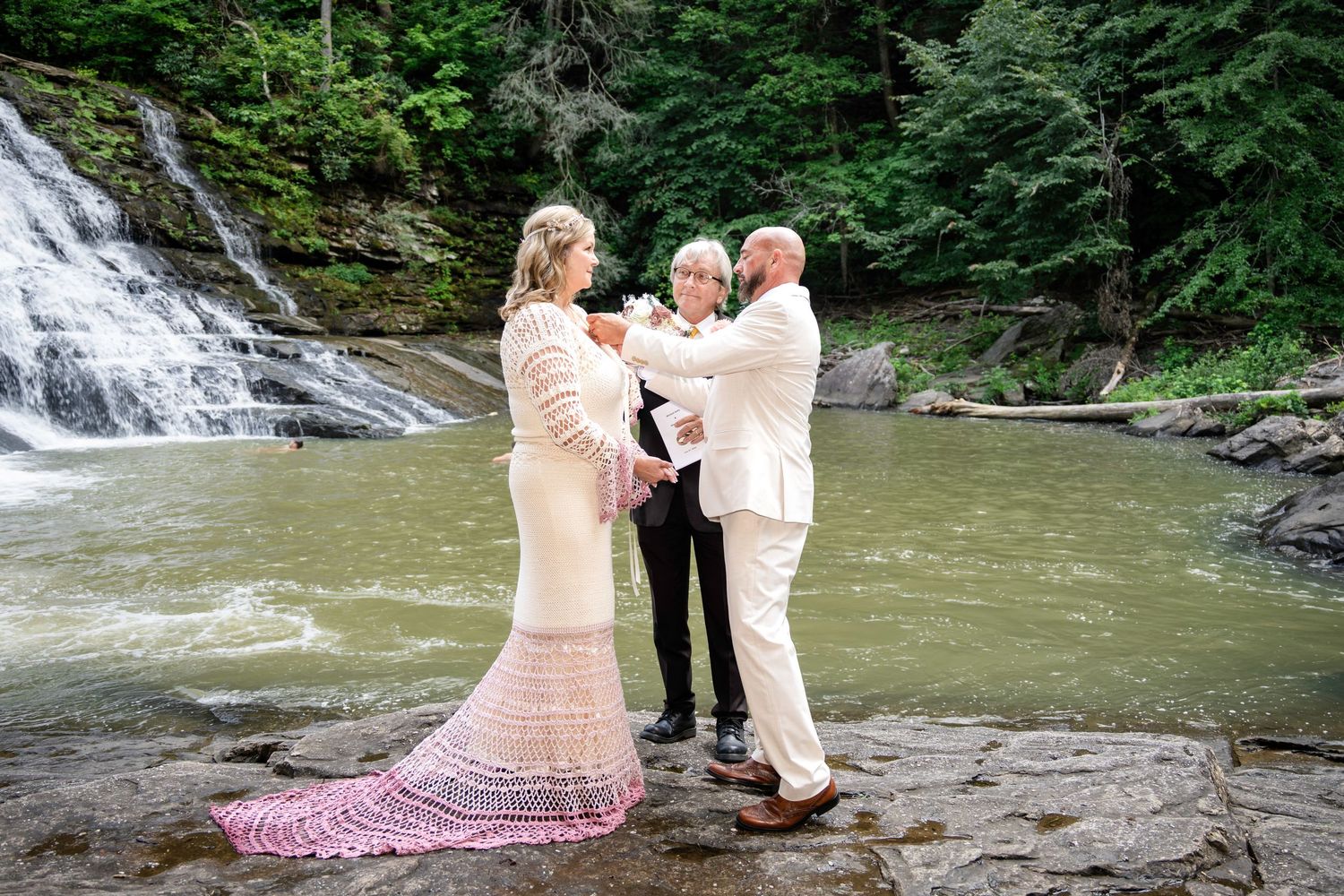 Couple exchanges vows with officiant by a scenic waterfall on rocky terrain, bride in lace gown with pink shawl.