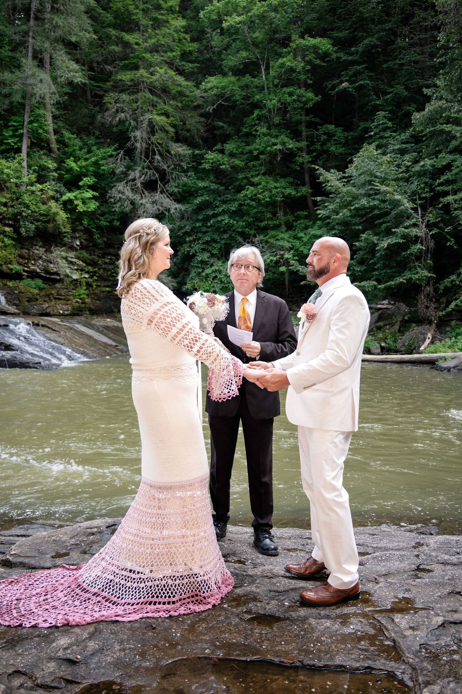 Bride and groom exchange vows outdoors by a waterfall with an officiant, surrounded by lush green forest.