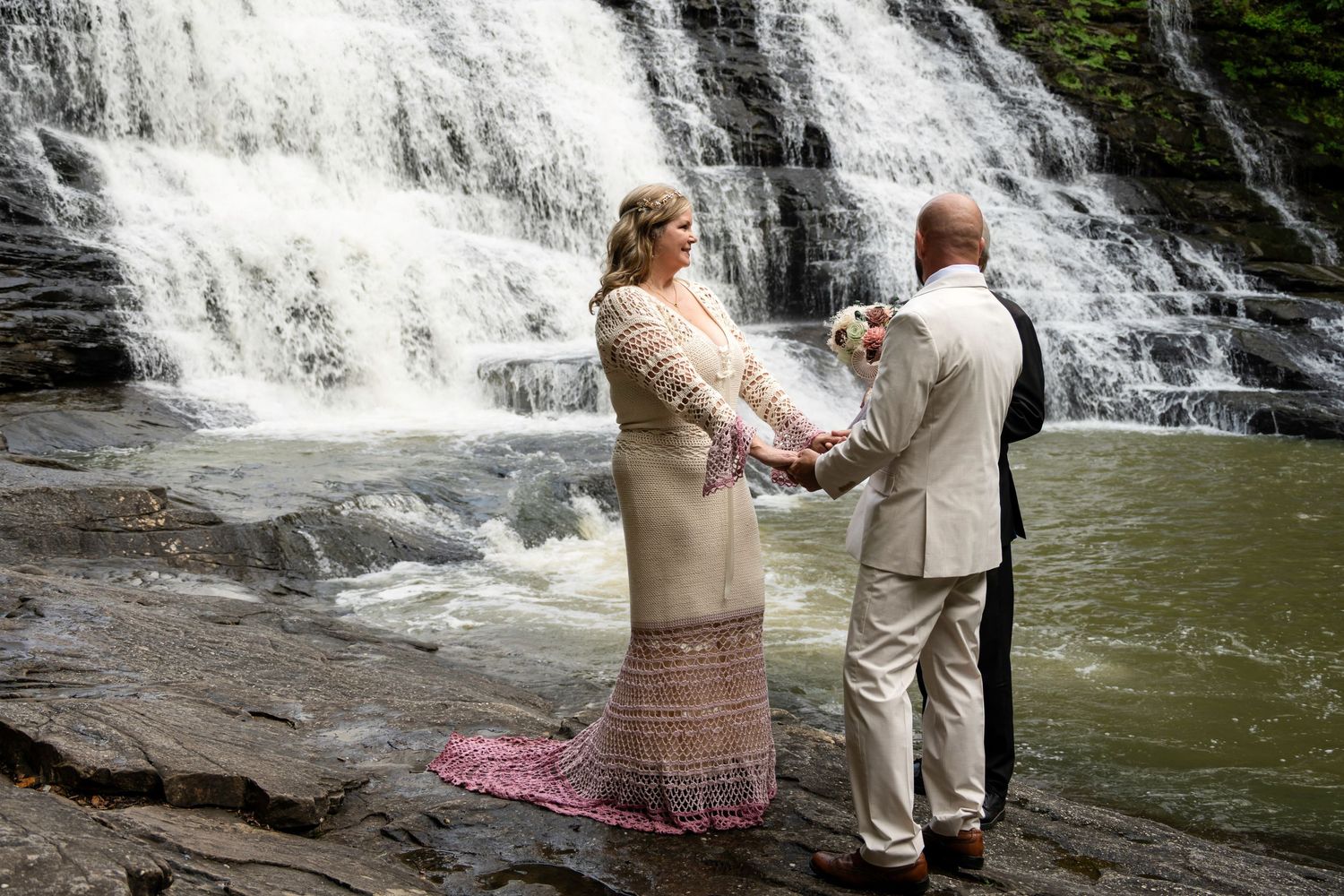 Couple exchanging vows in front of a stunning waterfall, bride in lace gown holding bouquet, groom in tan suit.