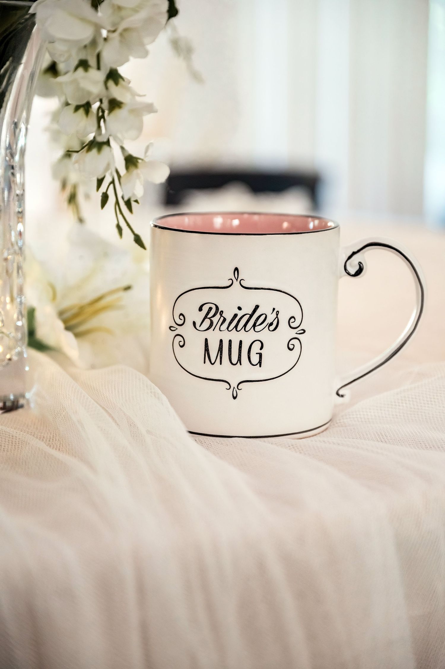 White ceramic Bride's Mug with pink interior and decorative black script, displayed on white fabric near white flowers.