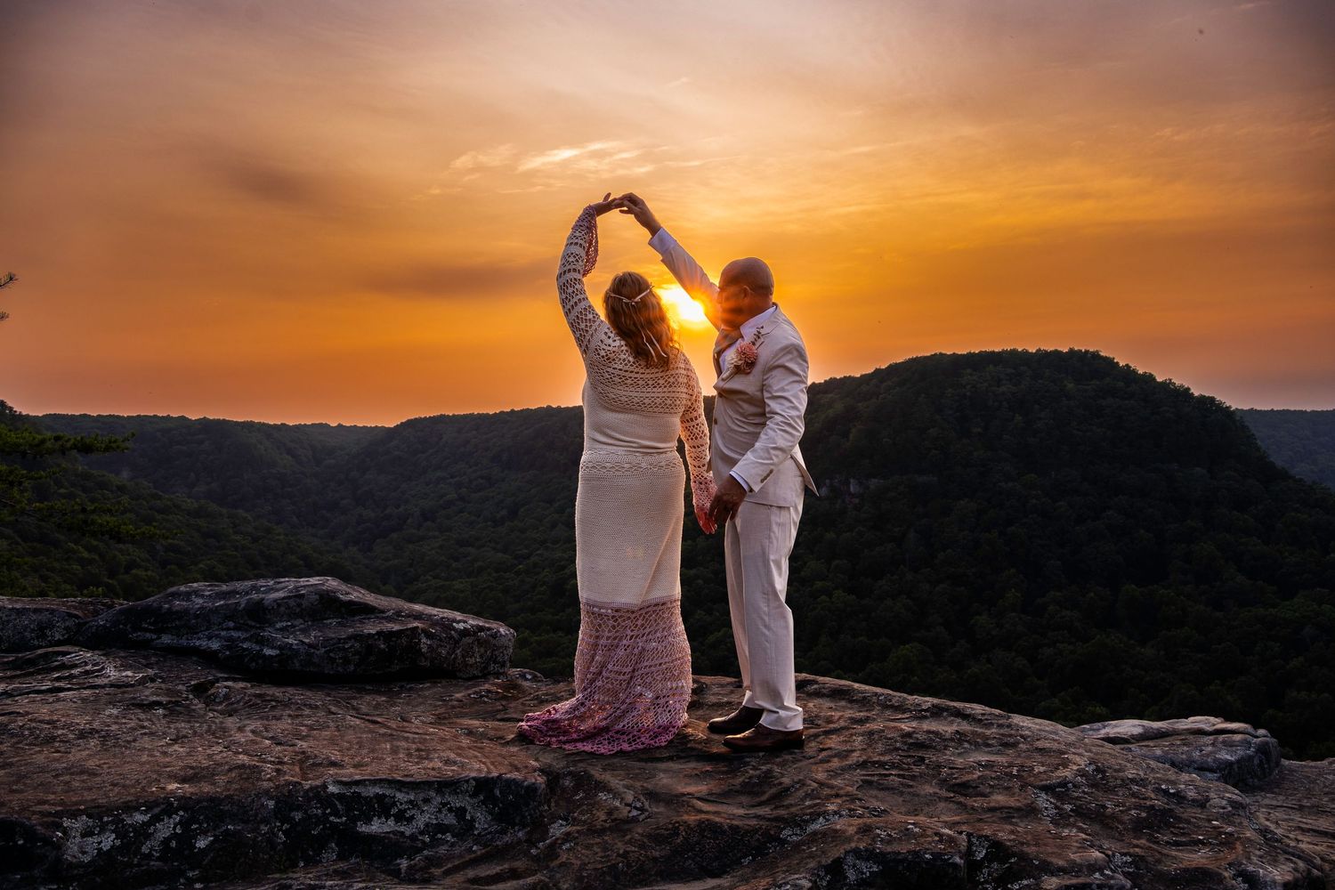 Couple holding hands at sunset on a rocky cliff overlooking a lush valley, forming a heart shape with their arms.