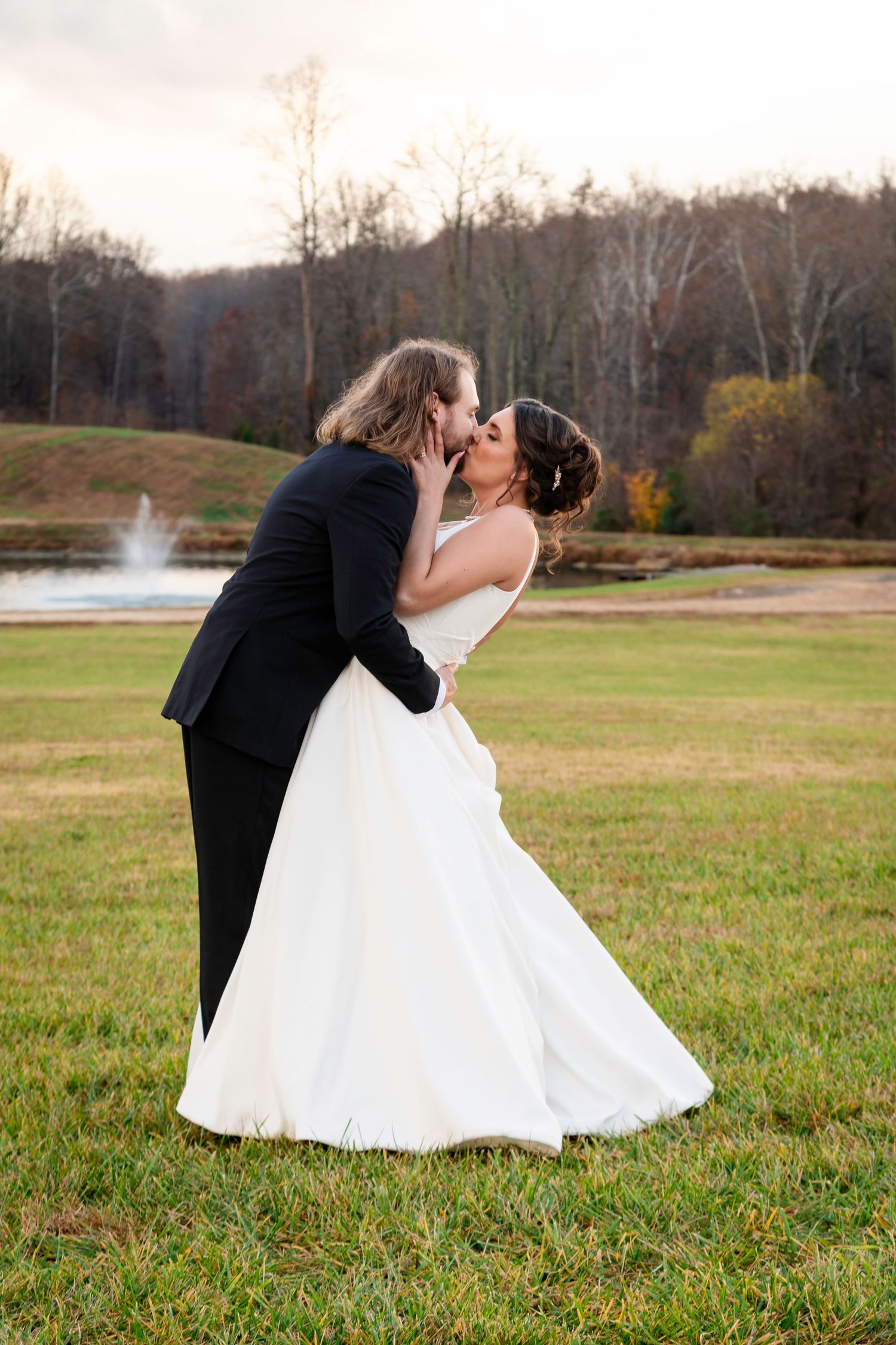 Bride and groom share a dip kiss on green lawn with fountain and autumn trees behind them.