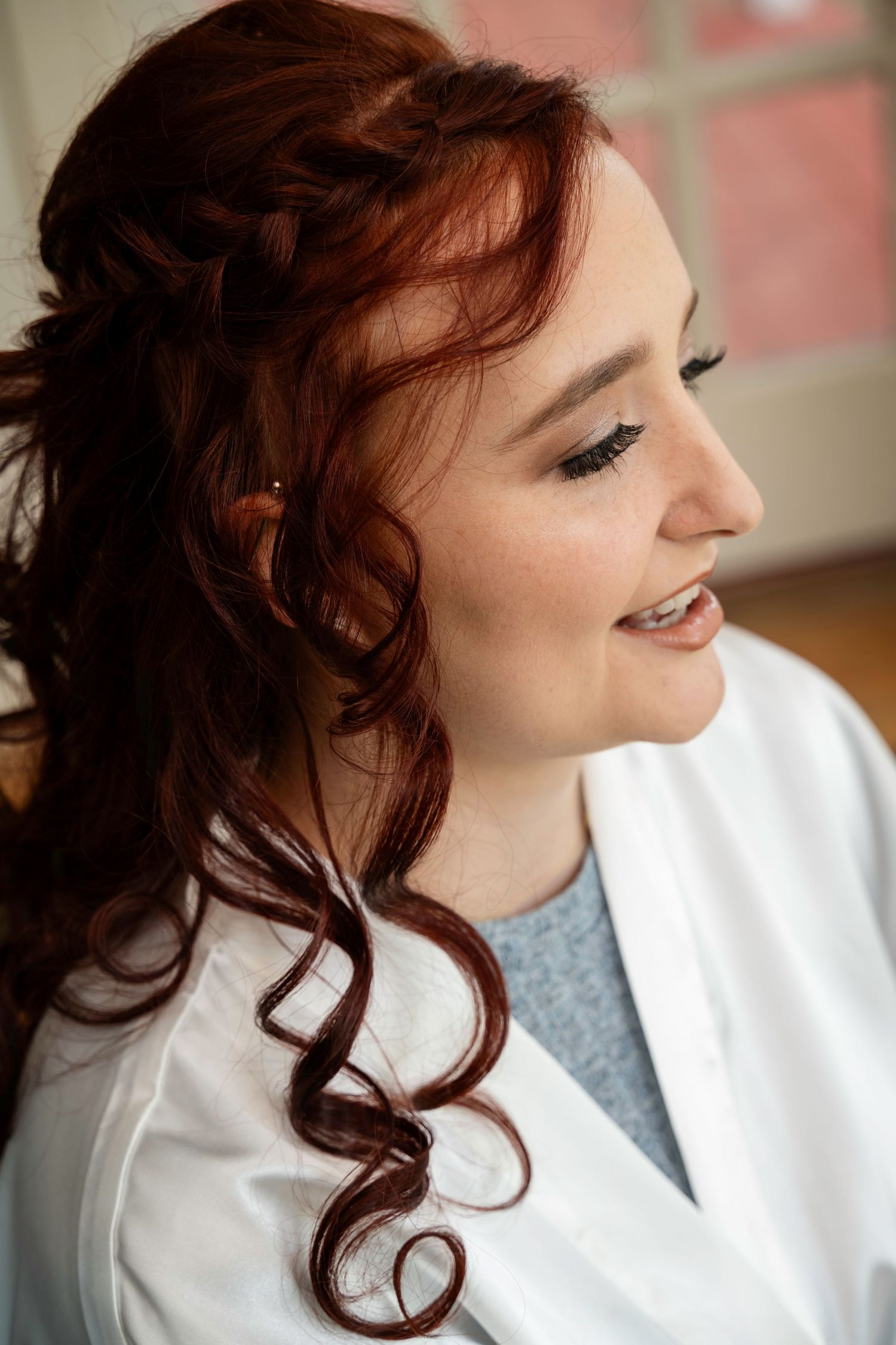 Close-up of bride with elegant red curly hair half-up style and natural bridal makeup wearing white robe.