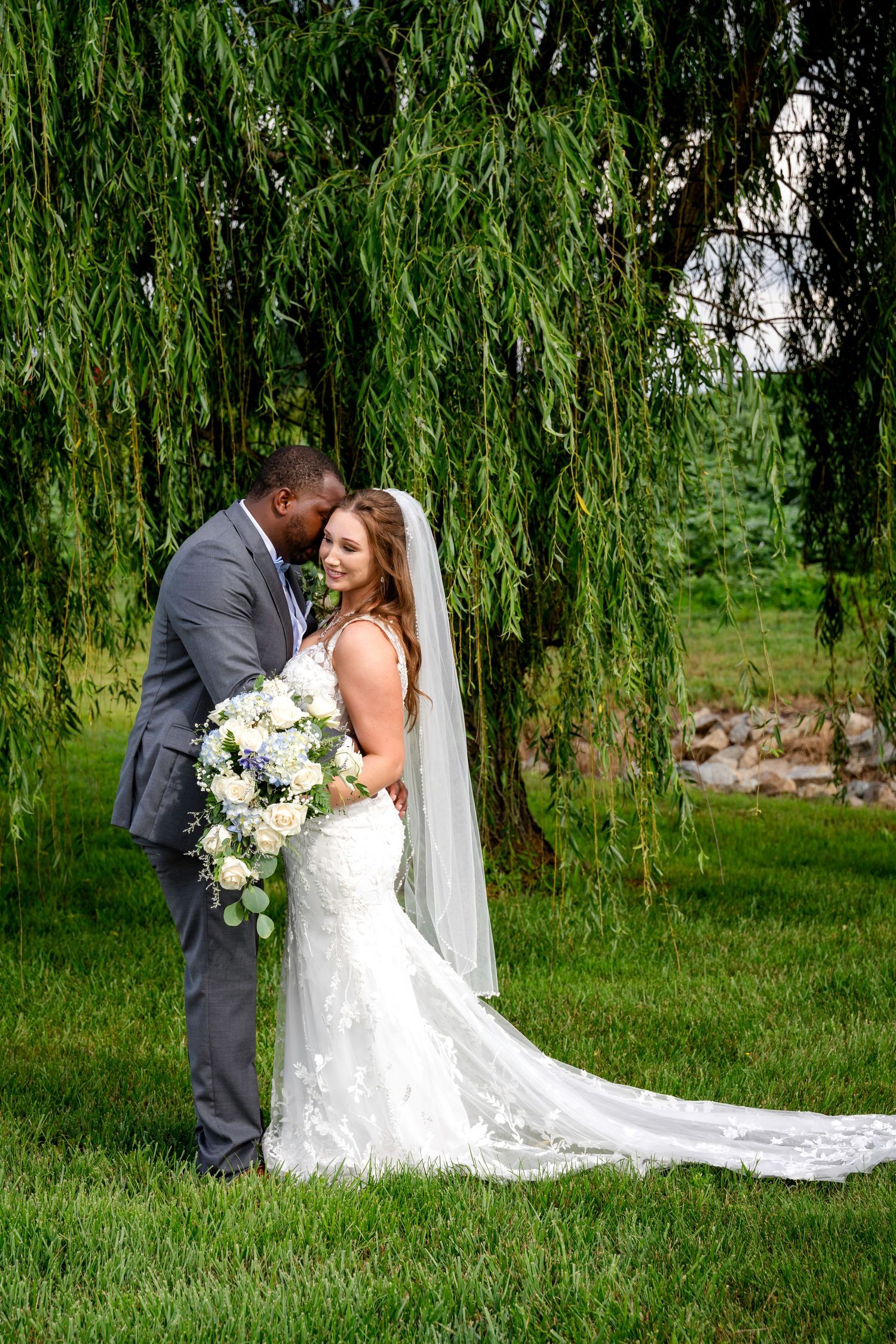 Bride and groom sharing tender moment under weeping willow tree, bride holding white and blue floral bouquet.