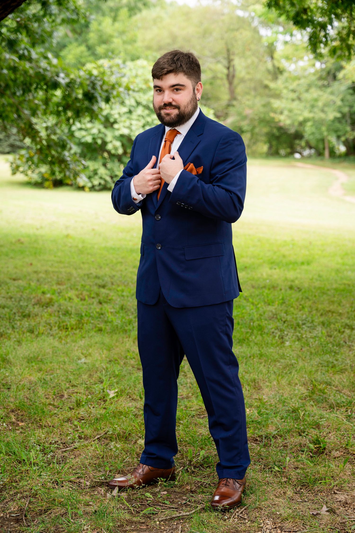 Dapper groom in a navy blue suit with orange tie posing outdoors in a park.