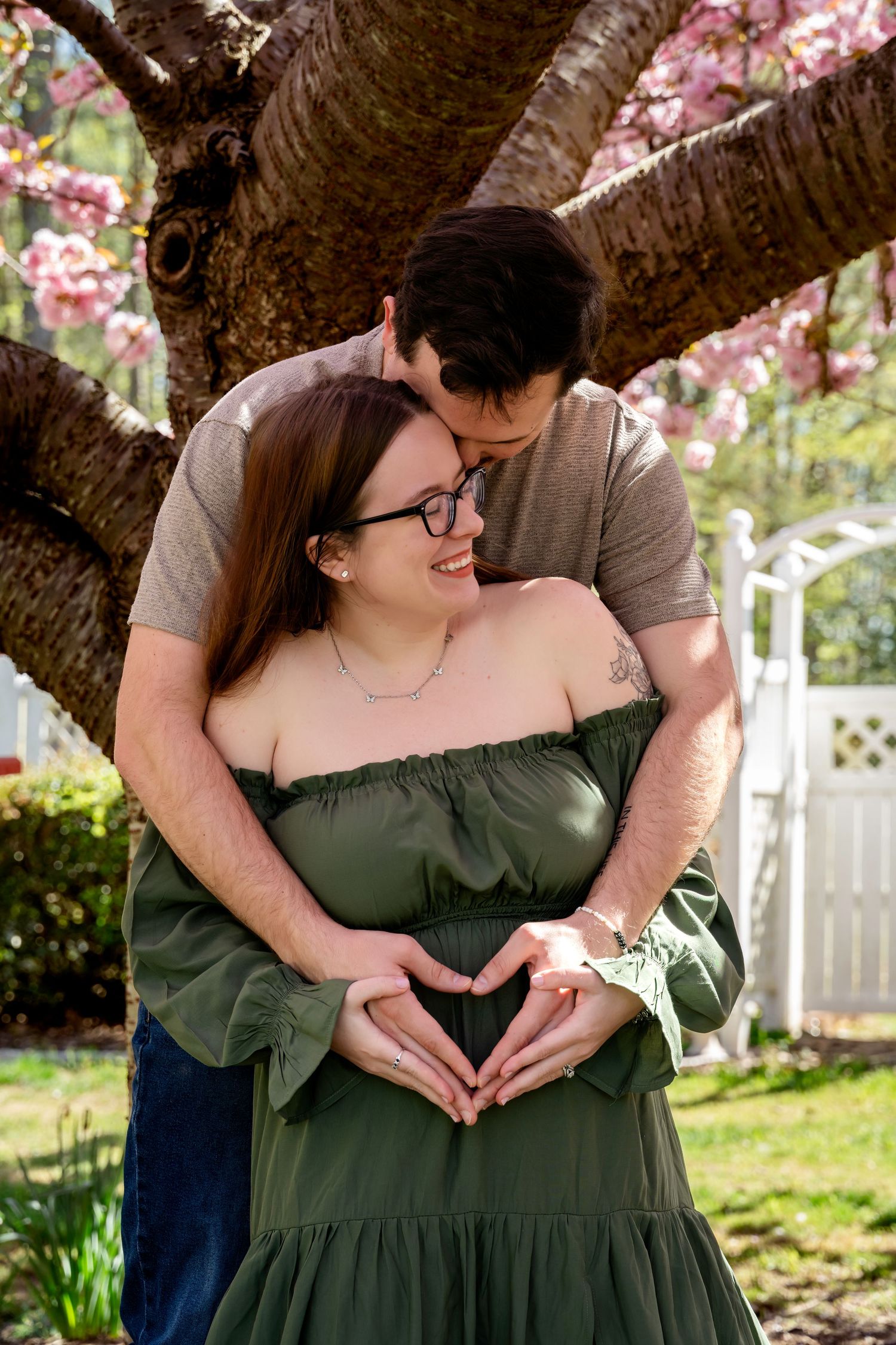 Couple sharing a tender maternity moment under blooming cherry tree, making heart shape over baby bump in green dress.