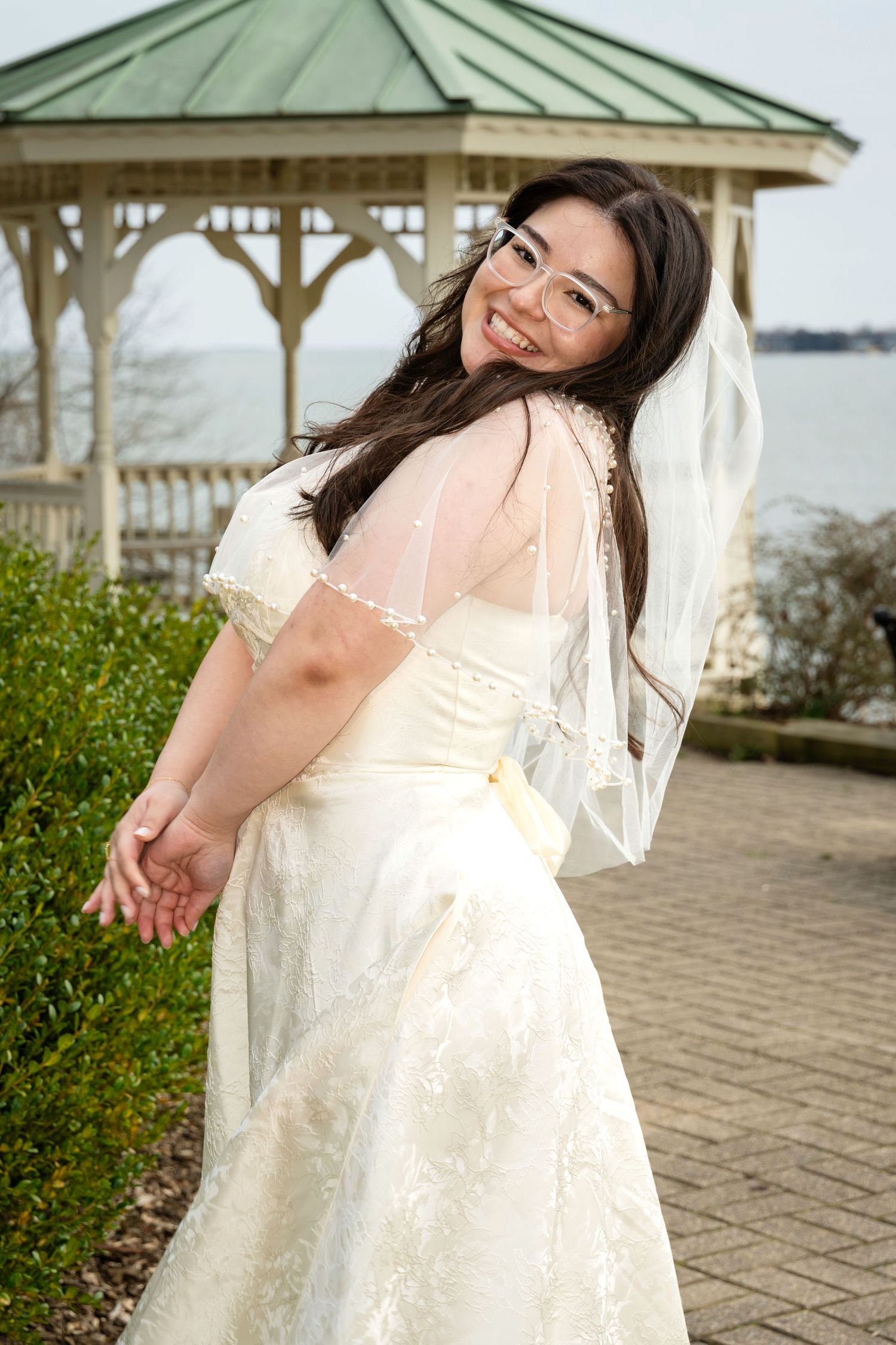Smiling bride in white lace gown and veil poses near gazebo by waterfront on her wedding day.