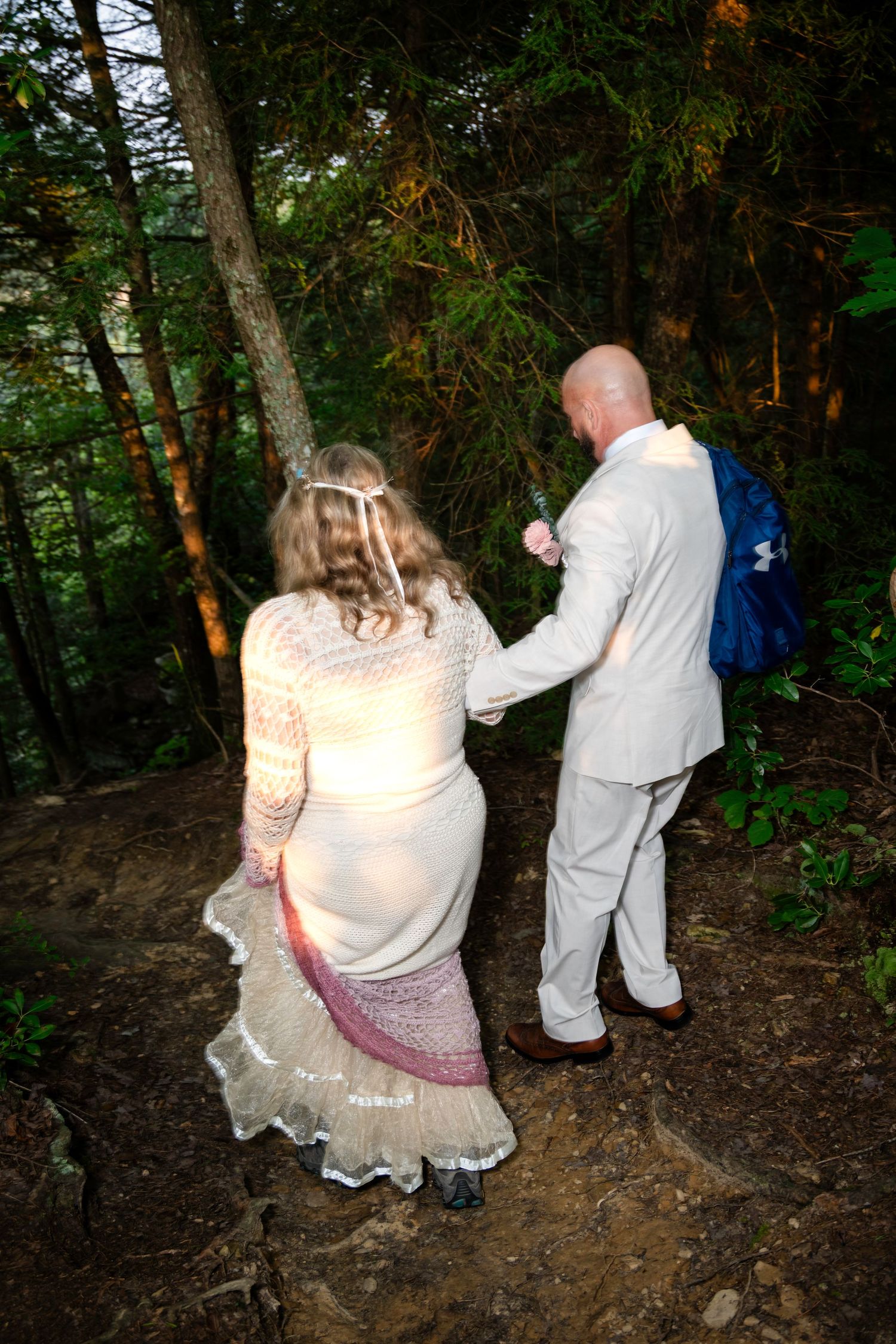 A bride and groom hold hands while walking through a lush forest trail during their outdoor woodland wedding ceremony.
