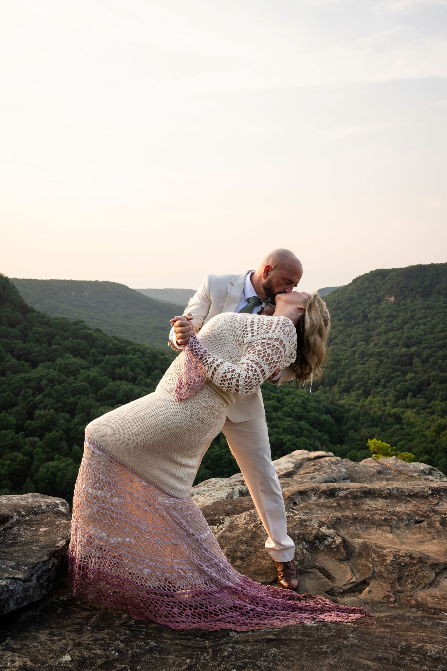 Bride and groom share romantic dip kiss on rocky mountain overlook with lush green valley backdrop at sunset.