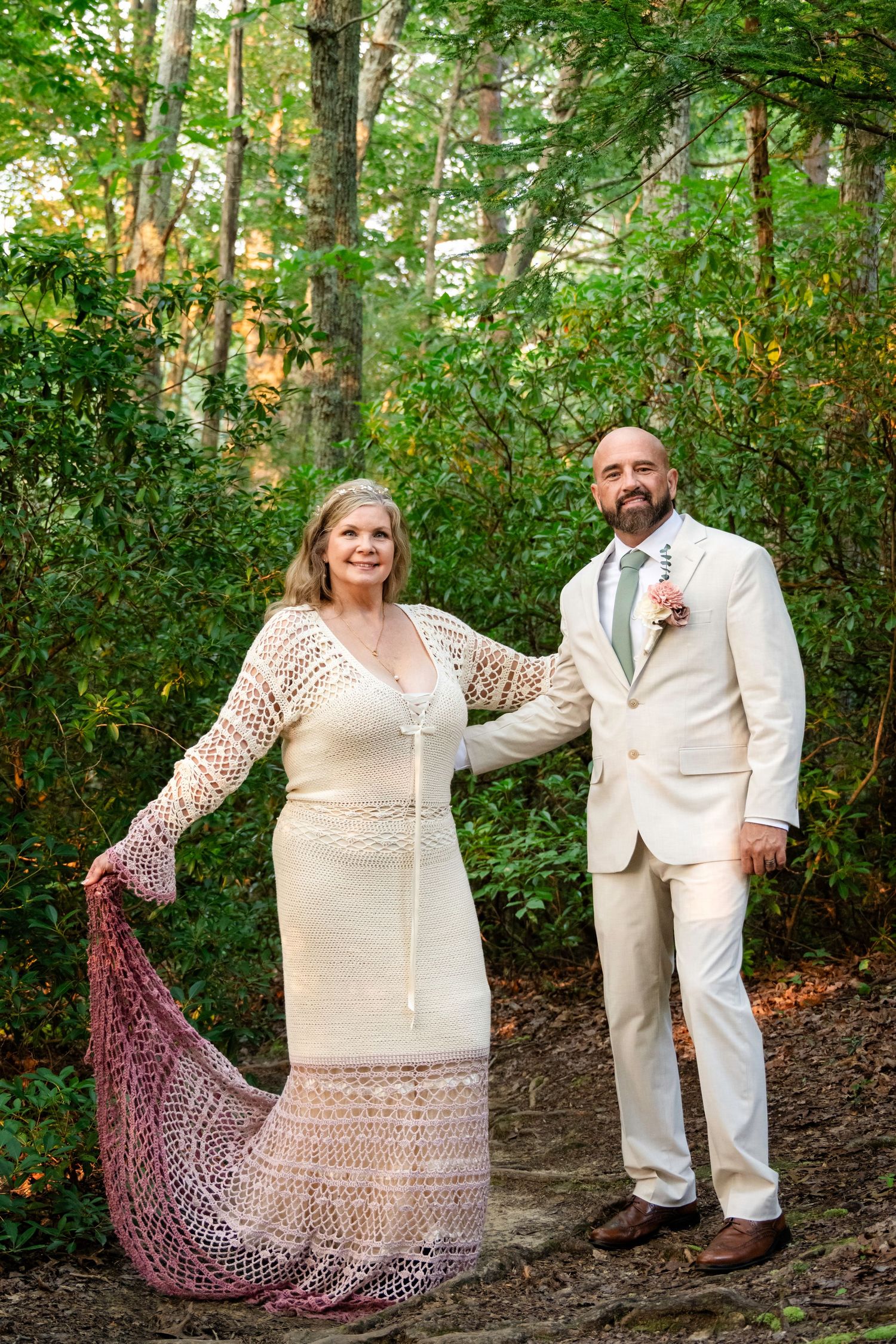 Couple in wedding attire posing in a lush forest, bride in lace dress with shawl, groom in cream suit with boutonniere.