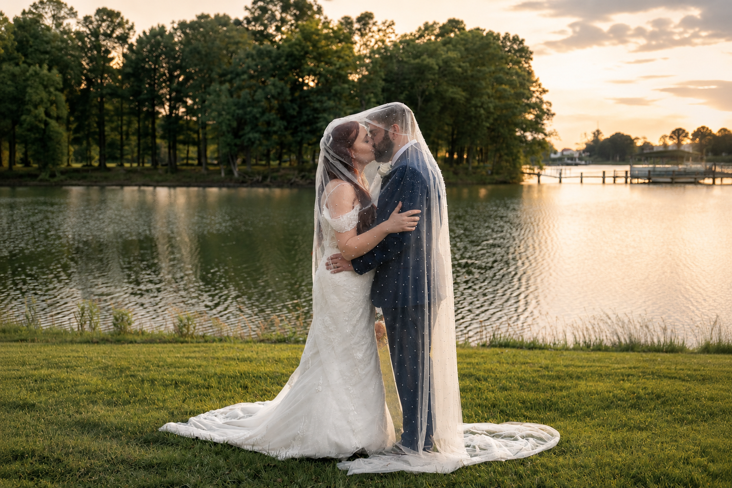 Bride and groom sharing a romantic kiss under a veil at sunset by a serene lake surrounded by lush trees.