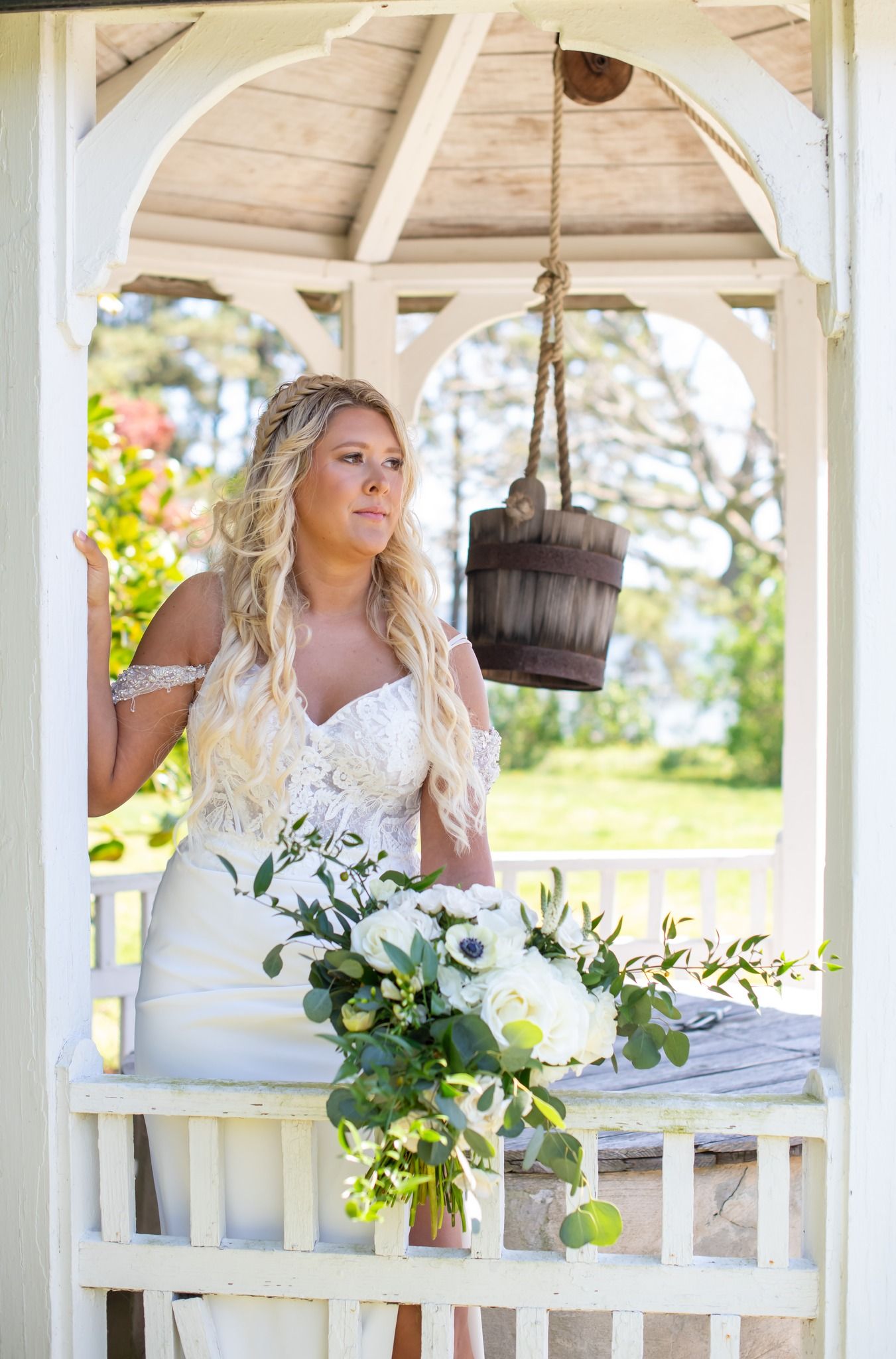 Blonde bride in off-shoulder lace gown holding bouquet inside rustic white gazebo.