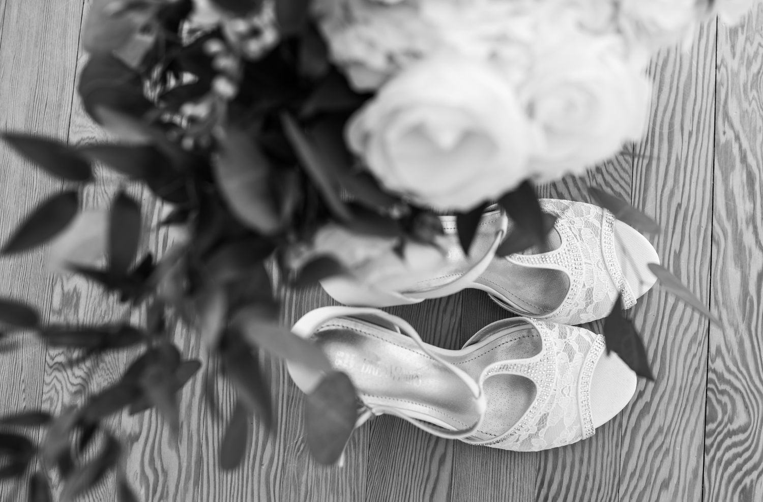 Black and white photo of lace bridal heels beside floral bouquet on wooden floor.