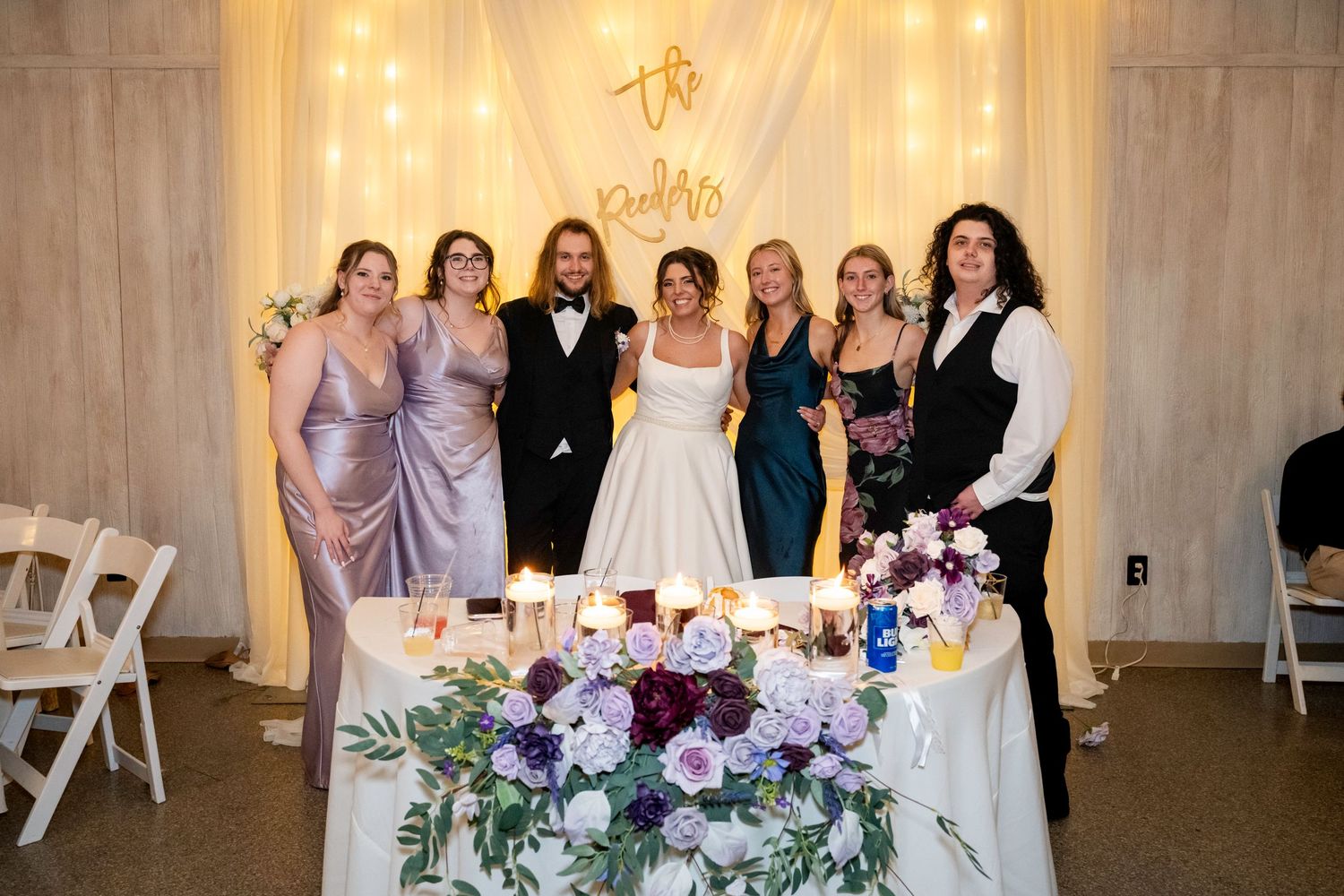 Bride and groom pose with bridesmaids at decorated wedding reception sweetheart table.