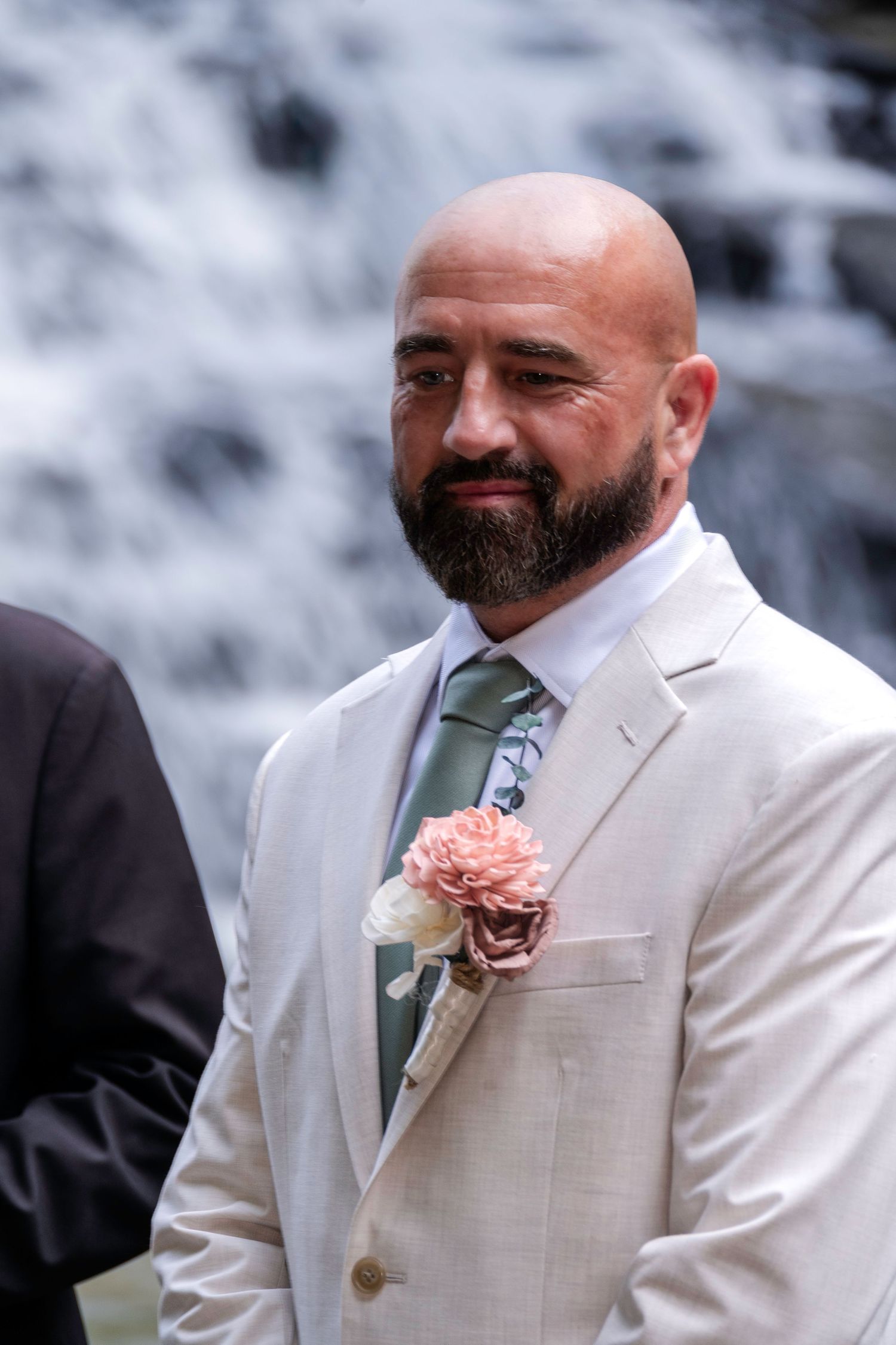 Bald groom with beard wearing ivory suit, floral boutonniere, and sage tie stands before waterfall backdrop.