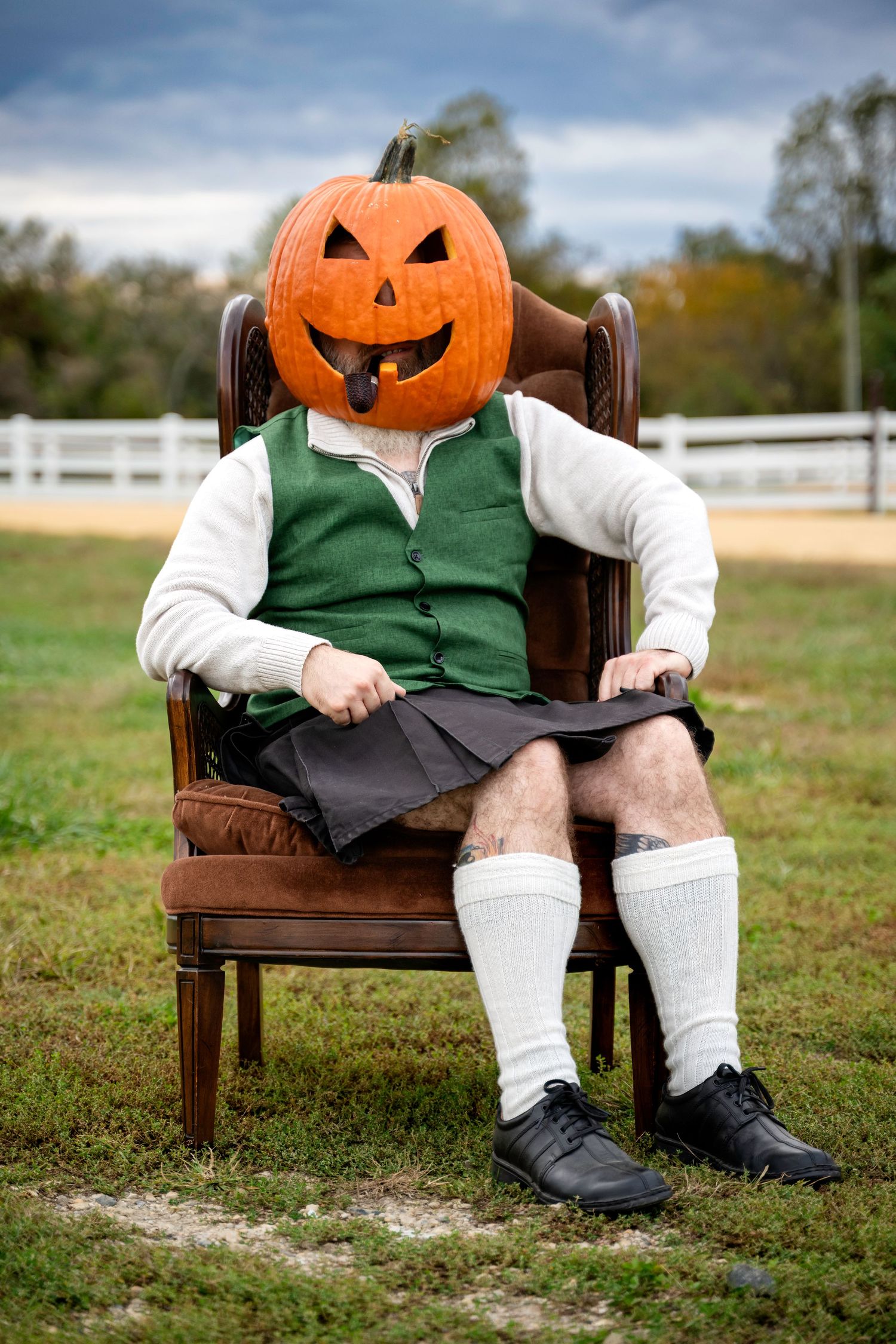Person wearing a jack-o-lantern pumpkin head sitting in an armchair outdoors in a green vest and kilt.