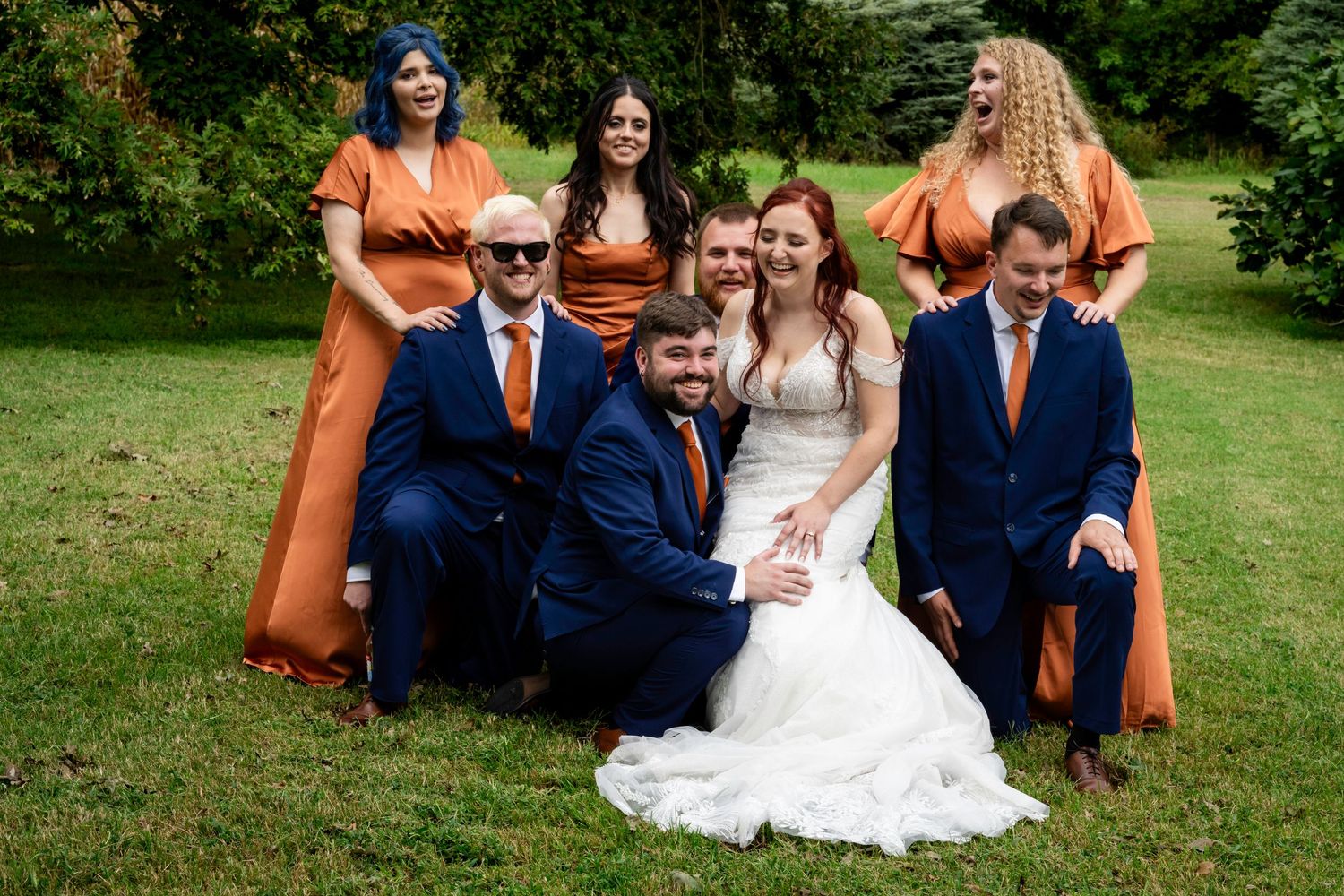 Wedding party in orange and navy poses playfully on green lawn with bride and groom.