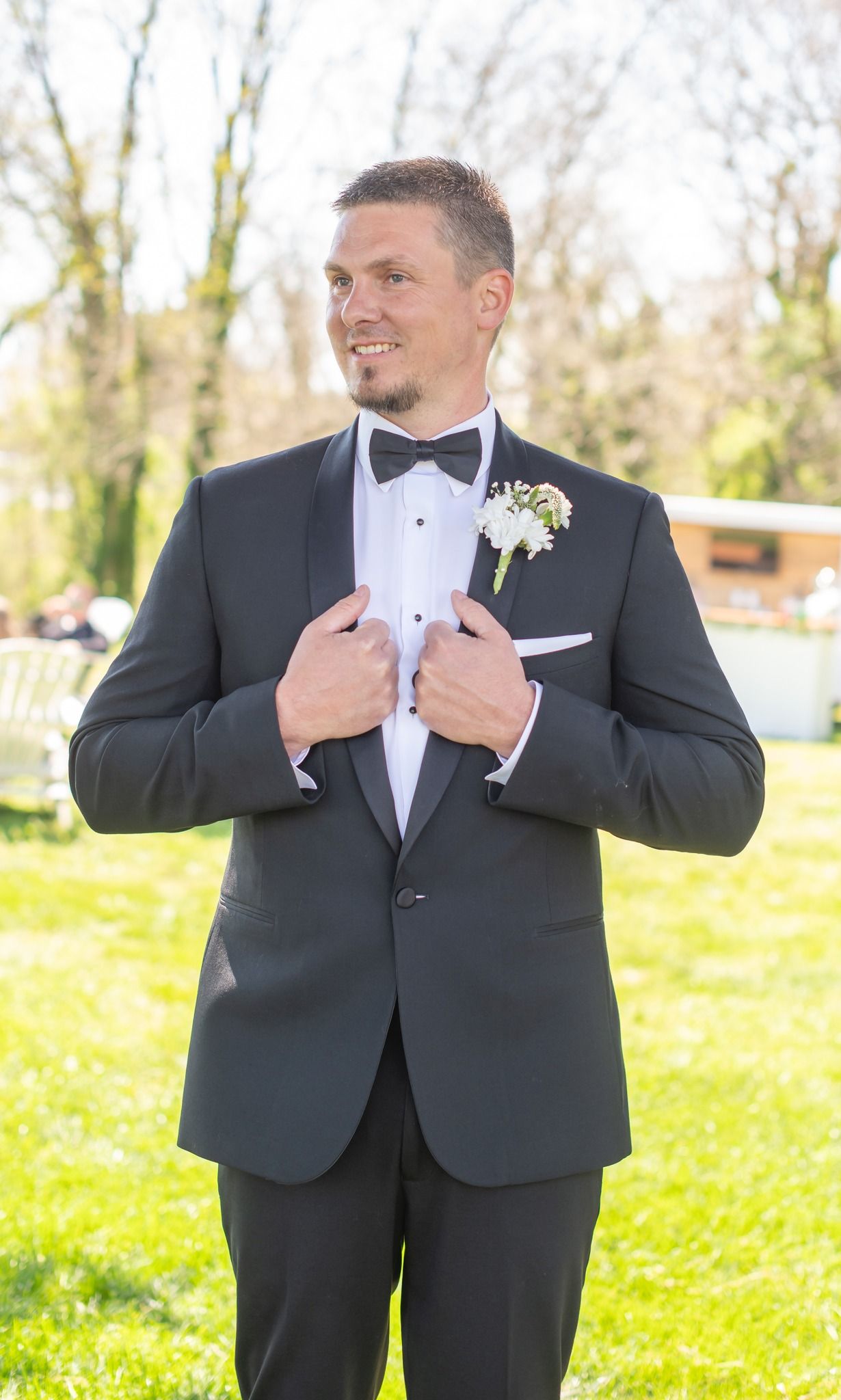 Groom in black tuxedo with bow tie and boutonniere posing outdoors on green grass.