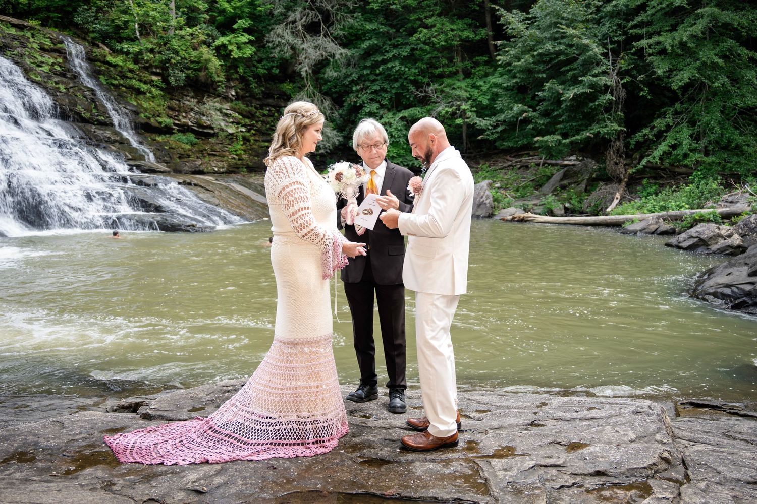 Couple exchanging vows with officiant beside a waterfall and river in a lush green forest outdoor wedding ceremony.