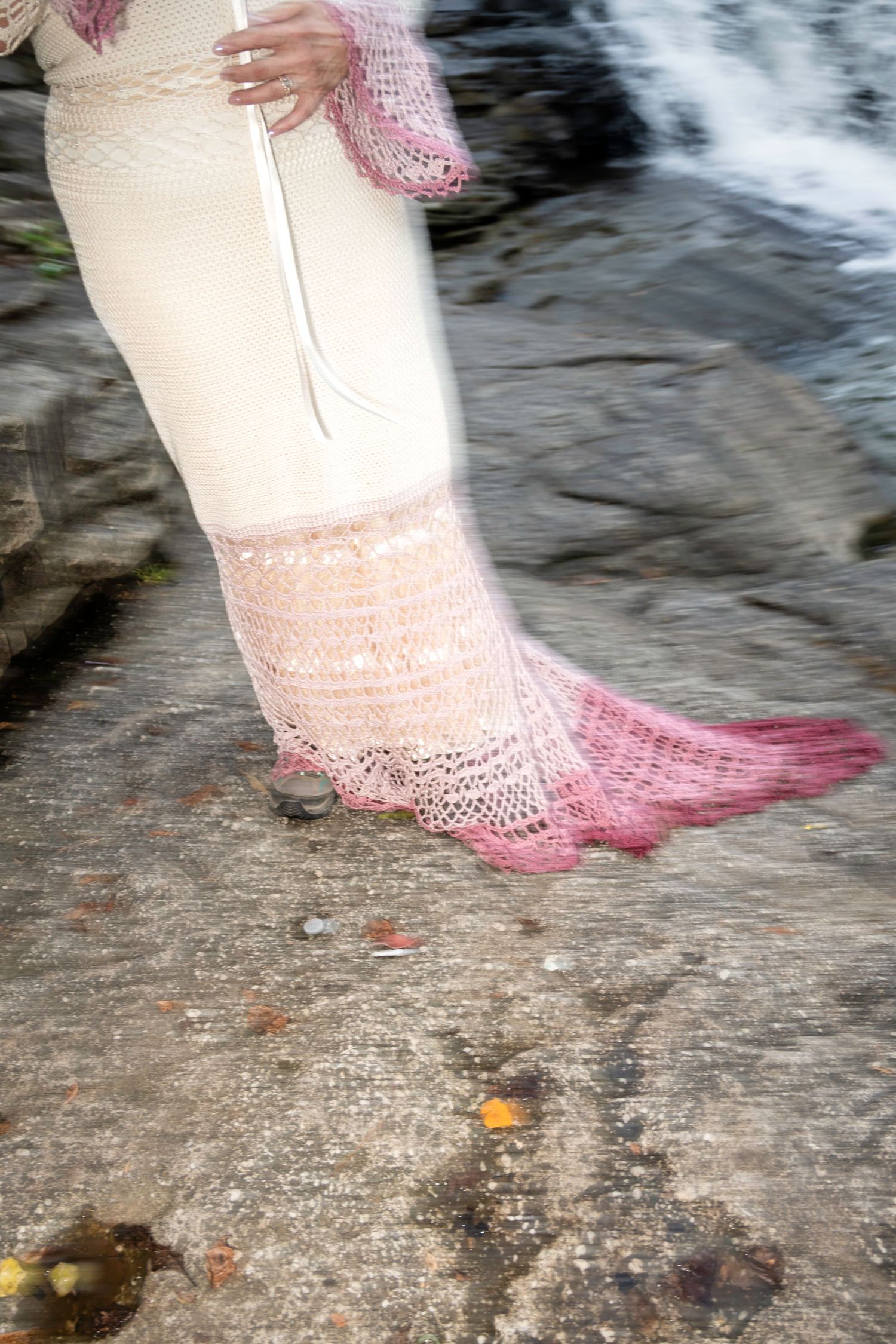 Person wearing a white and pink crochet lace skirt with pink trim standing on rocky ground near water.