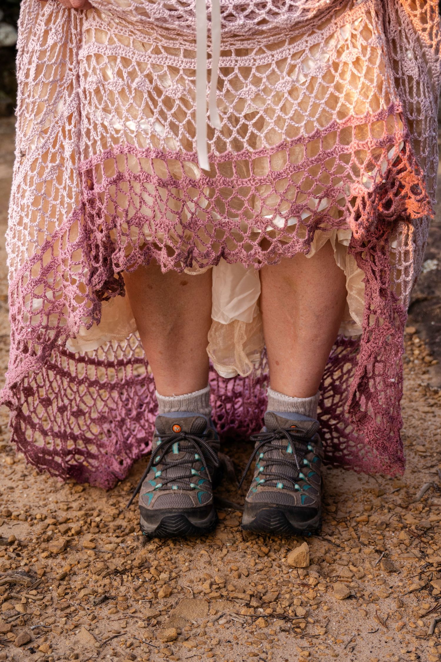 Close-up of bride's crochet ombre wedding dress hem with hiking shoes and socks visible underneath on rocky ground.