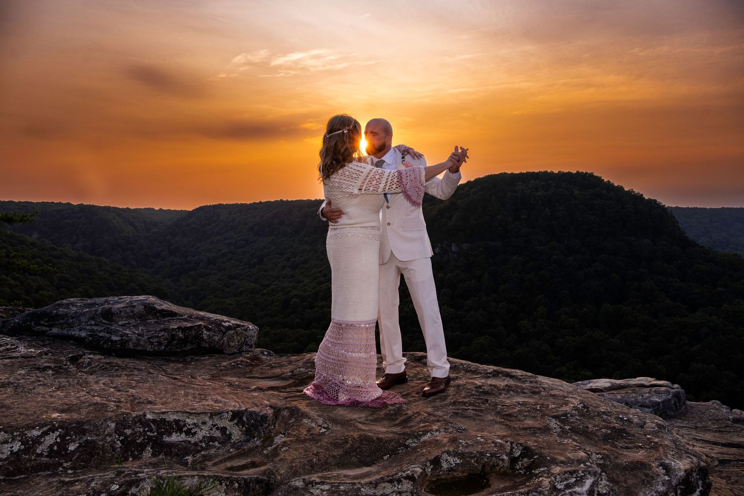 Couple dancing on rocky cliff at sunset with golden sky and lush mountain valley backdrop.