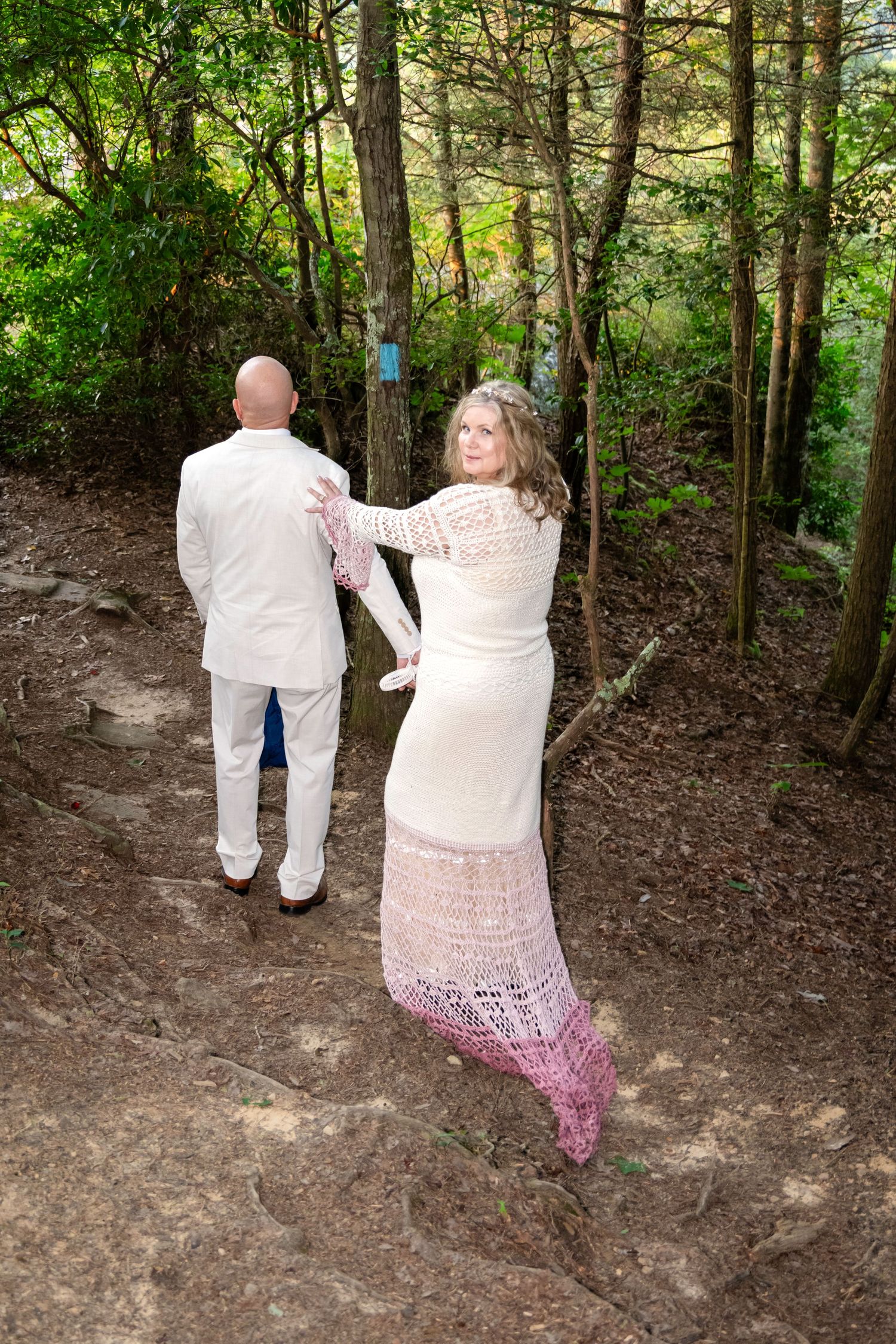 Bride in white lace gown and groom in white suit walk along a forest trail surrounded by lush green trees.