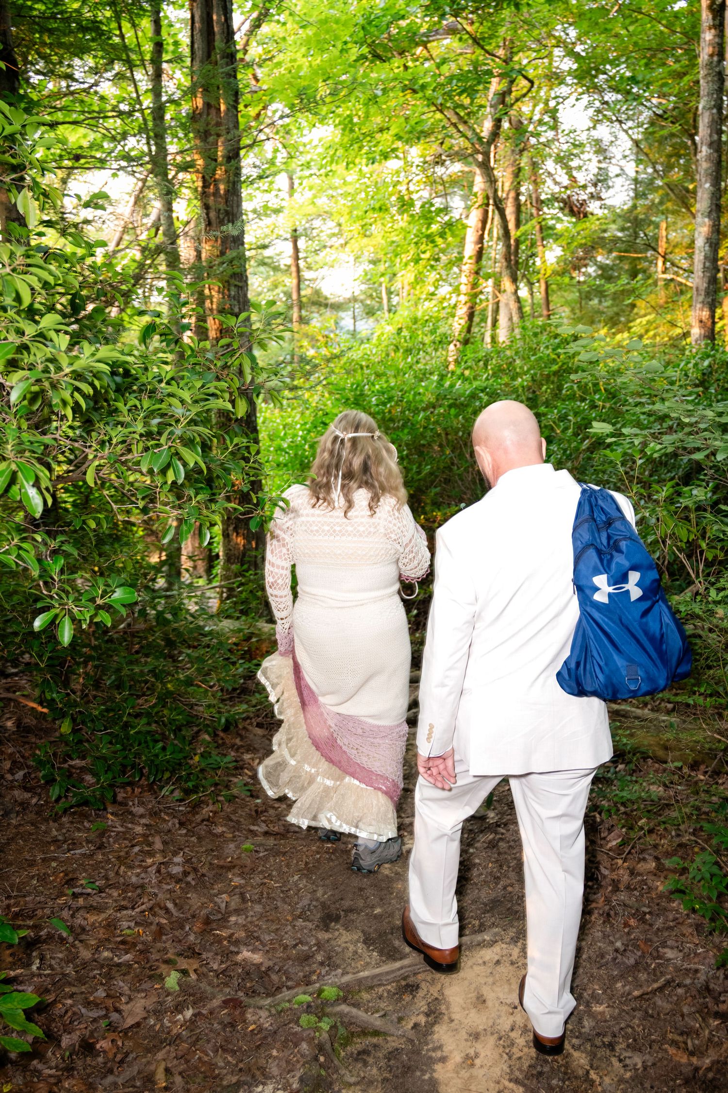 Bride and groom in white wedding attire walk hand-in-hand along a lush forest trail, man carrying blue backpack.
