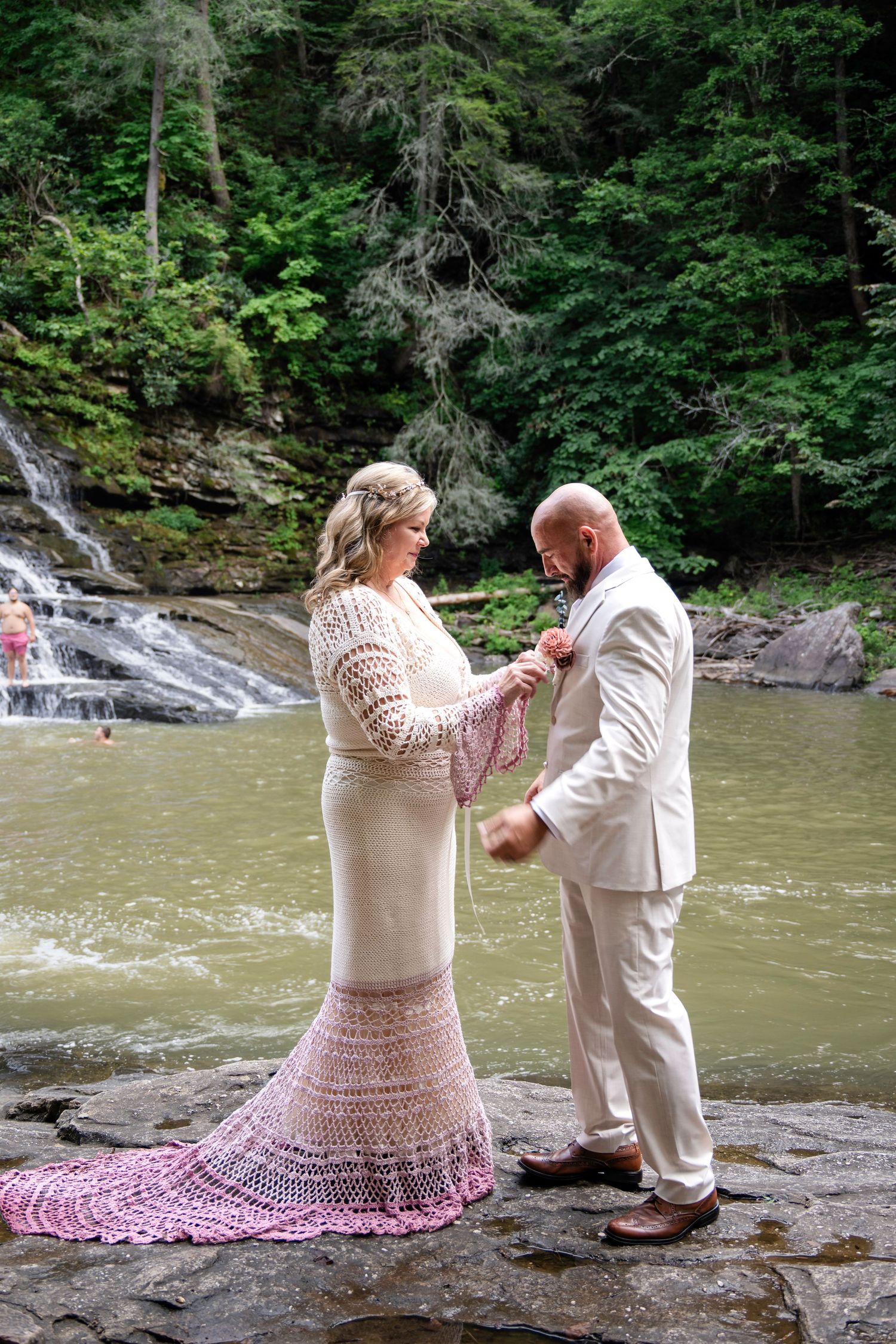 Bride in pink crochet gown pins boutonniere on groom in white suit beside a waterfall in lush forest.