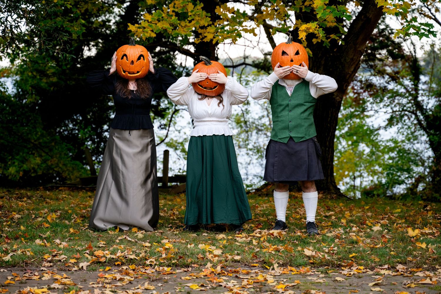 Three people in historical costumes with carved jack-o-lantern pumpkin heads pose outdoors among autumn fallen leaves.