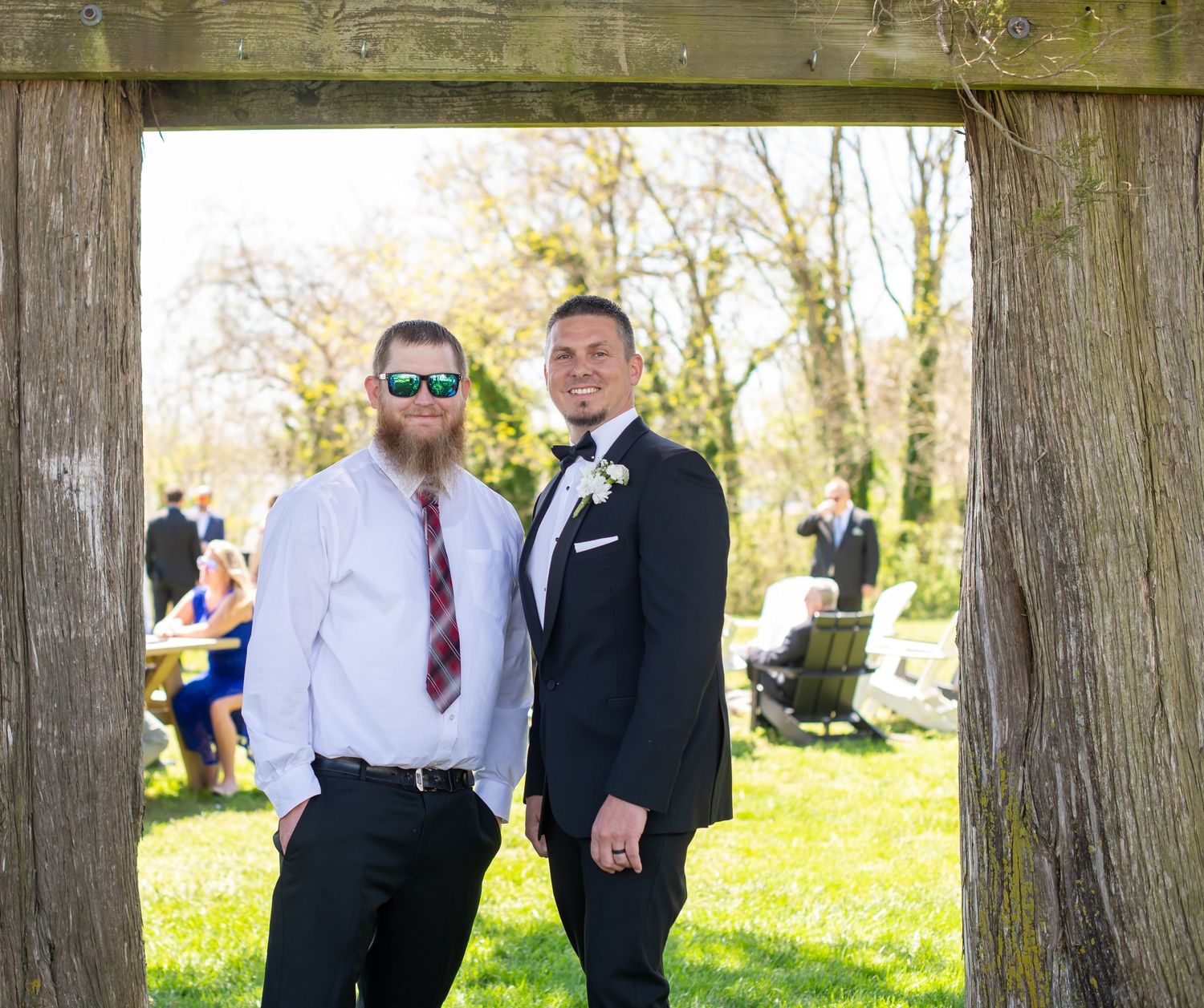 Groom and groomsman pose together under a rustic wooden arch at an outdoor wedding.
