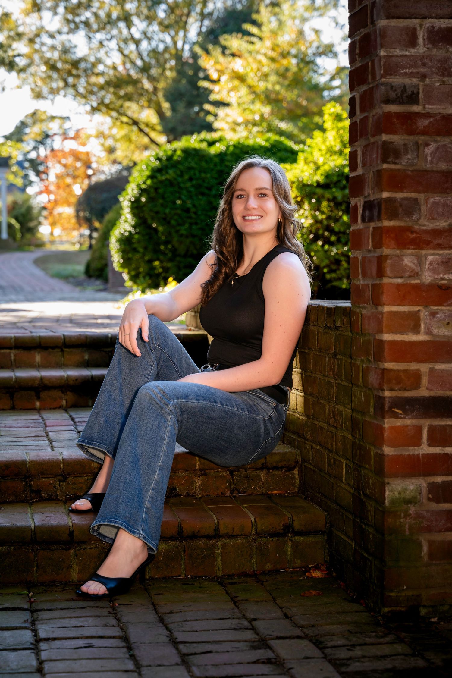 Smiling young woman sits on brick steps outdoors in black top and jeans on sunny day.