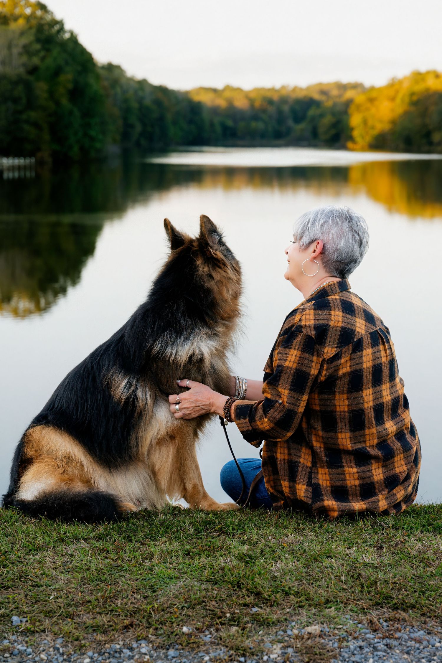 Older woman with short gray hair sitting by a calm lake with her German Shepherd dog at sunset.